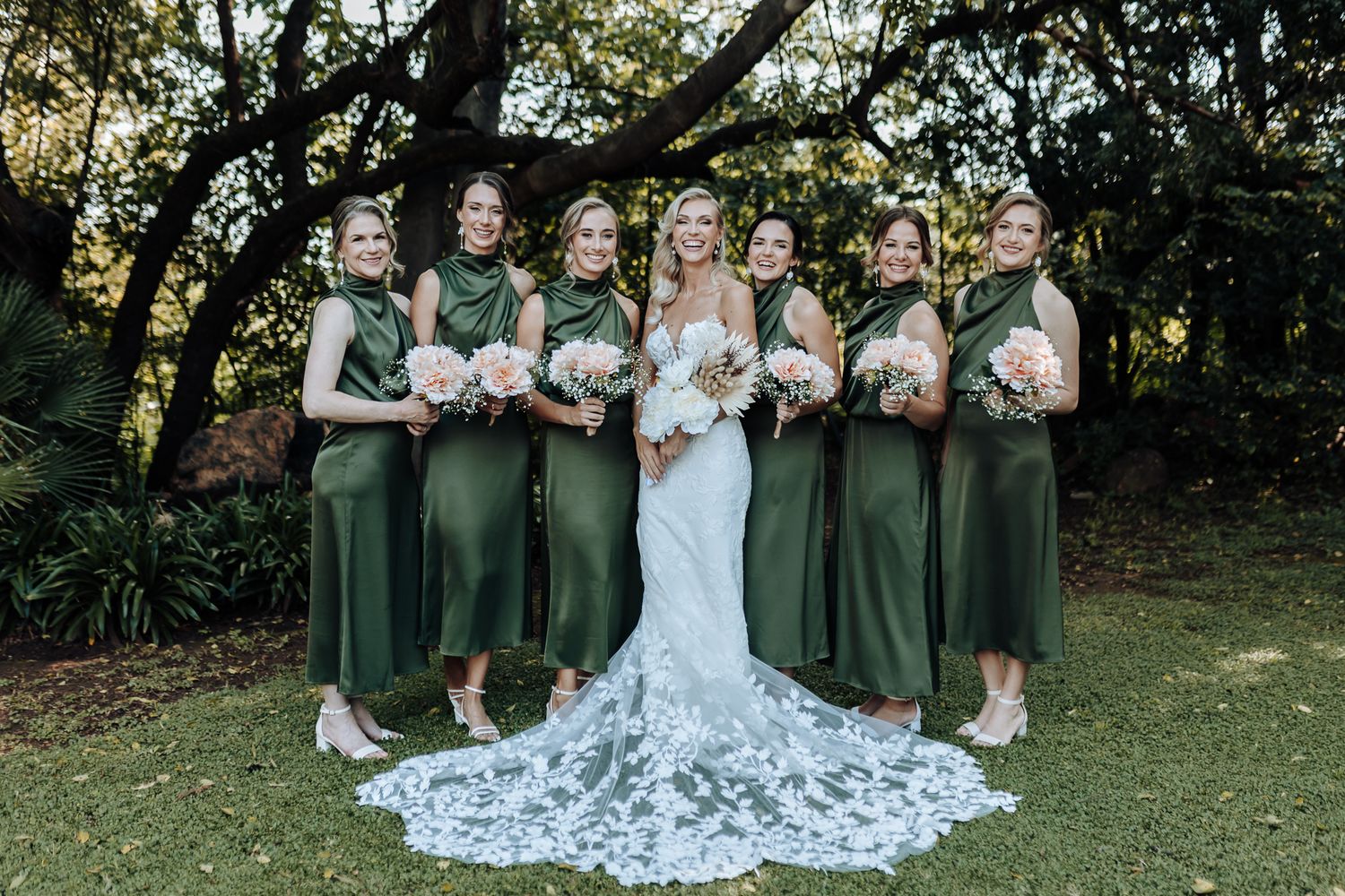 Wedding party in emerald dresses poses outdoors with stunning lace train on display.