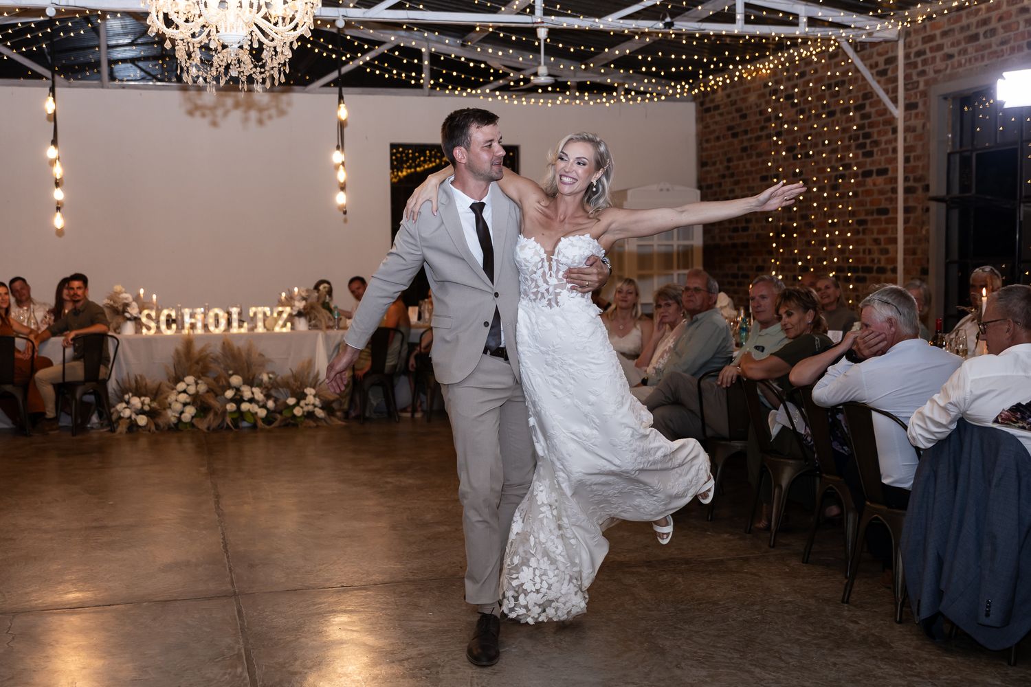 Couple performing energetic dance moves during wedding reception under chandeliers.