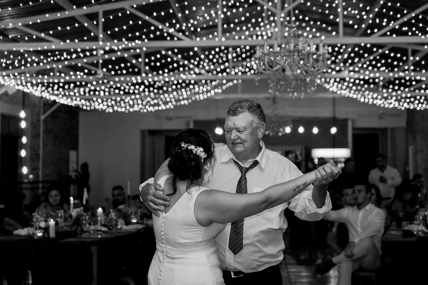 A black and white capture of a wedding dance beneath twinkling reception lights.