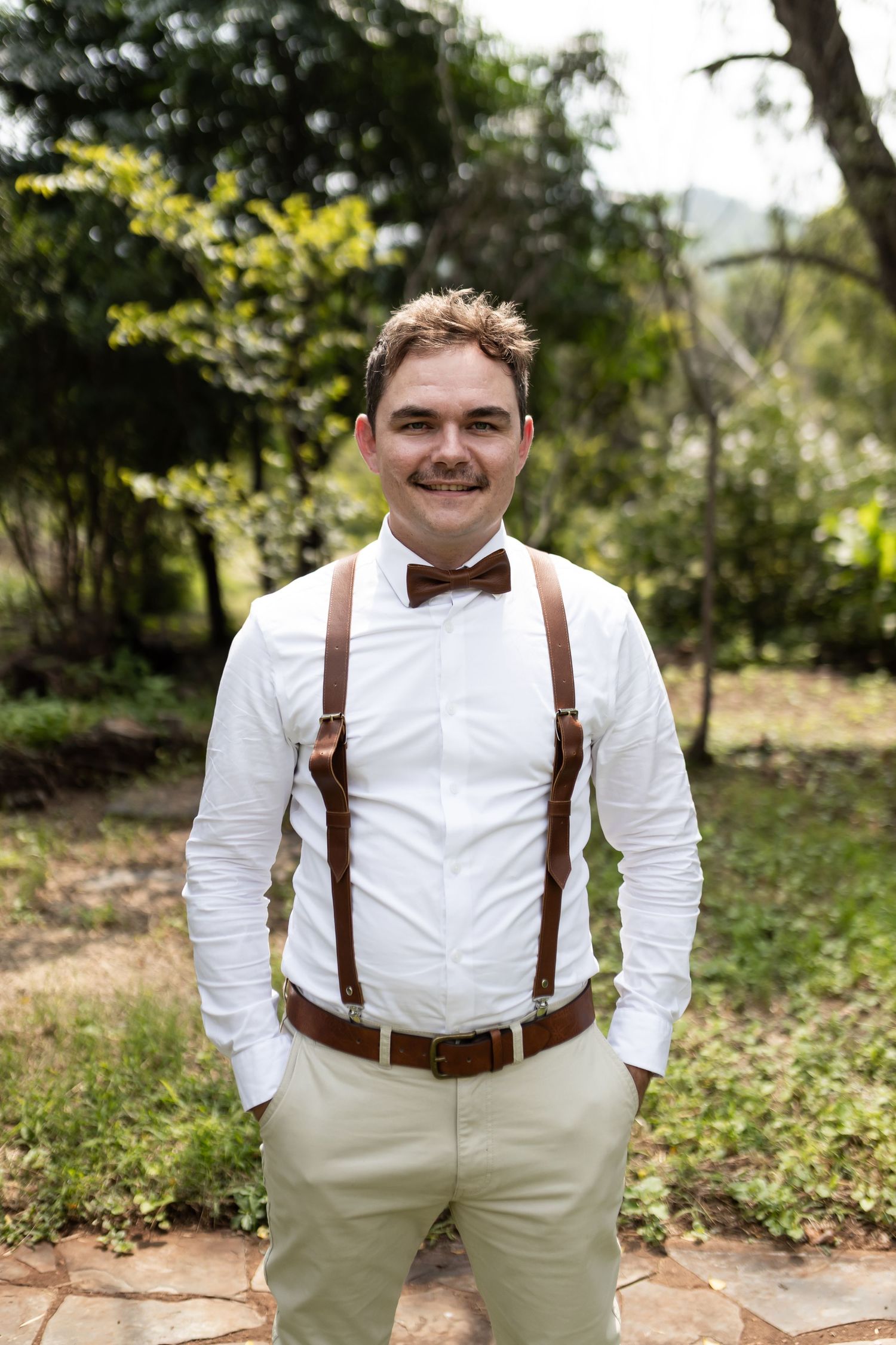 Groomsman in classic white shirt with brown suspenders poses in garden setting.