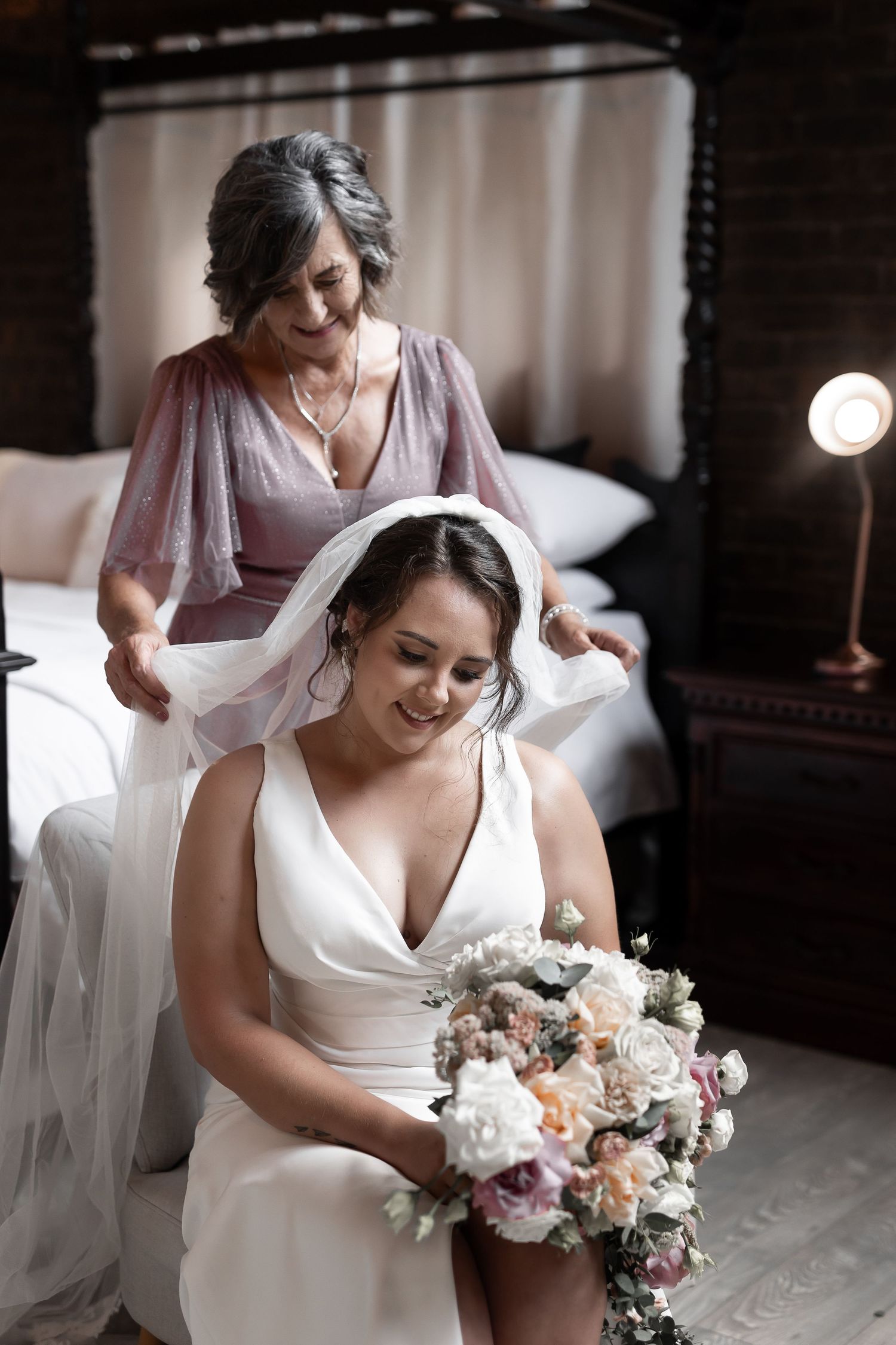 Wedding preparation scene with bride and helper adjusting veil in vintage bedroom setting.