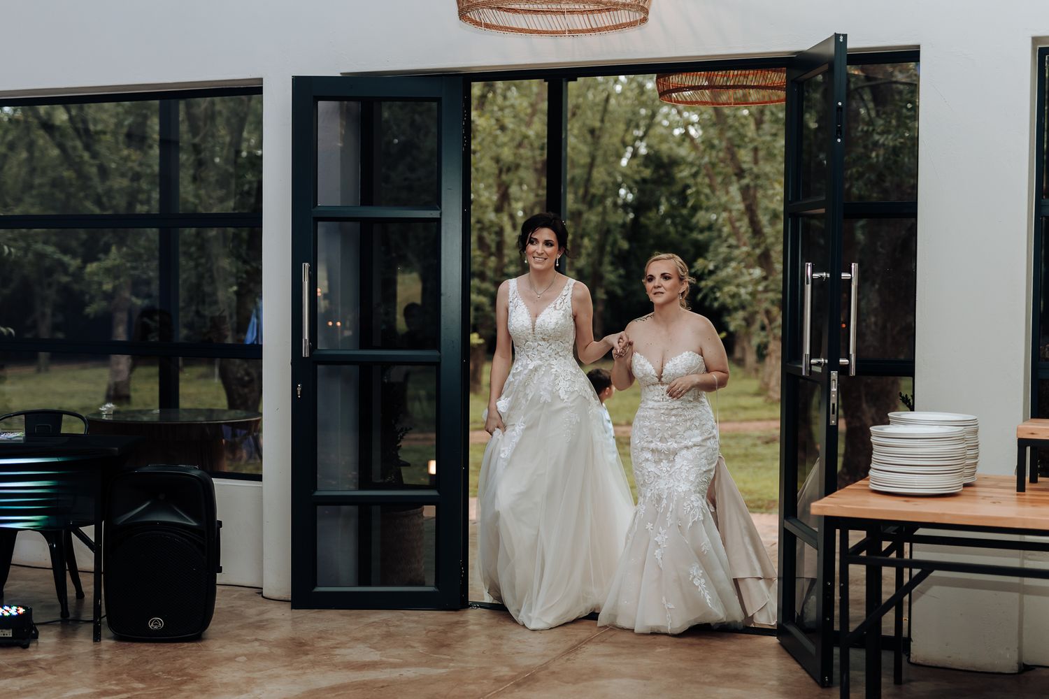 Two people in wedding gowns enter through black-framed glass doors at a modern venue.