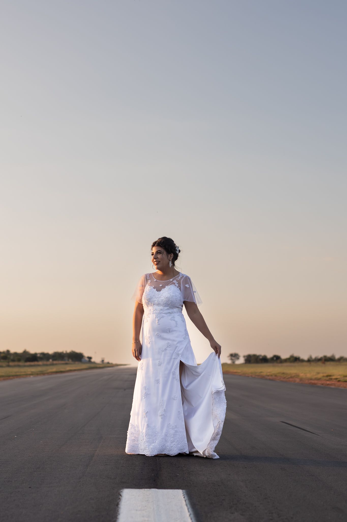 Bride in lace dress poses on empty runway during sunset.