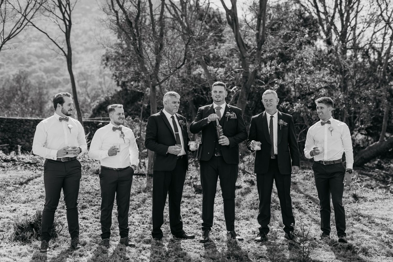 Wedding party dressed in dark suits stands together on rustic outdoor grounds.
