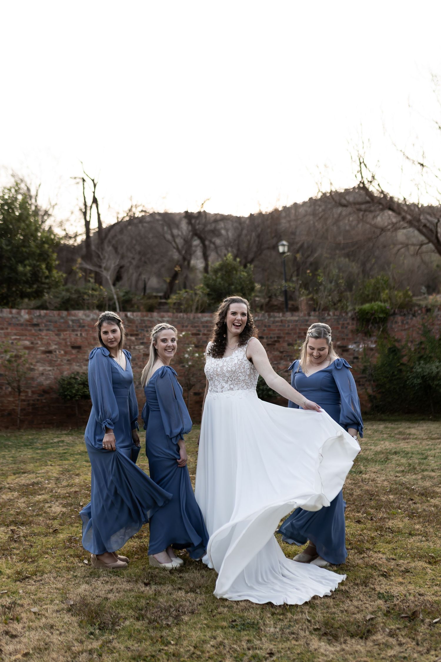 A group in matching blue dresses gathers around a white wedding dress flowing in the wind during a photo shoot outdoors.
