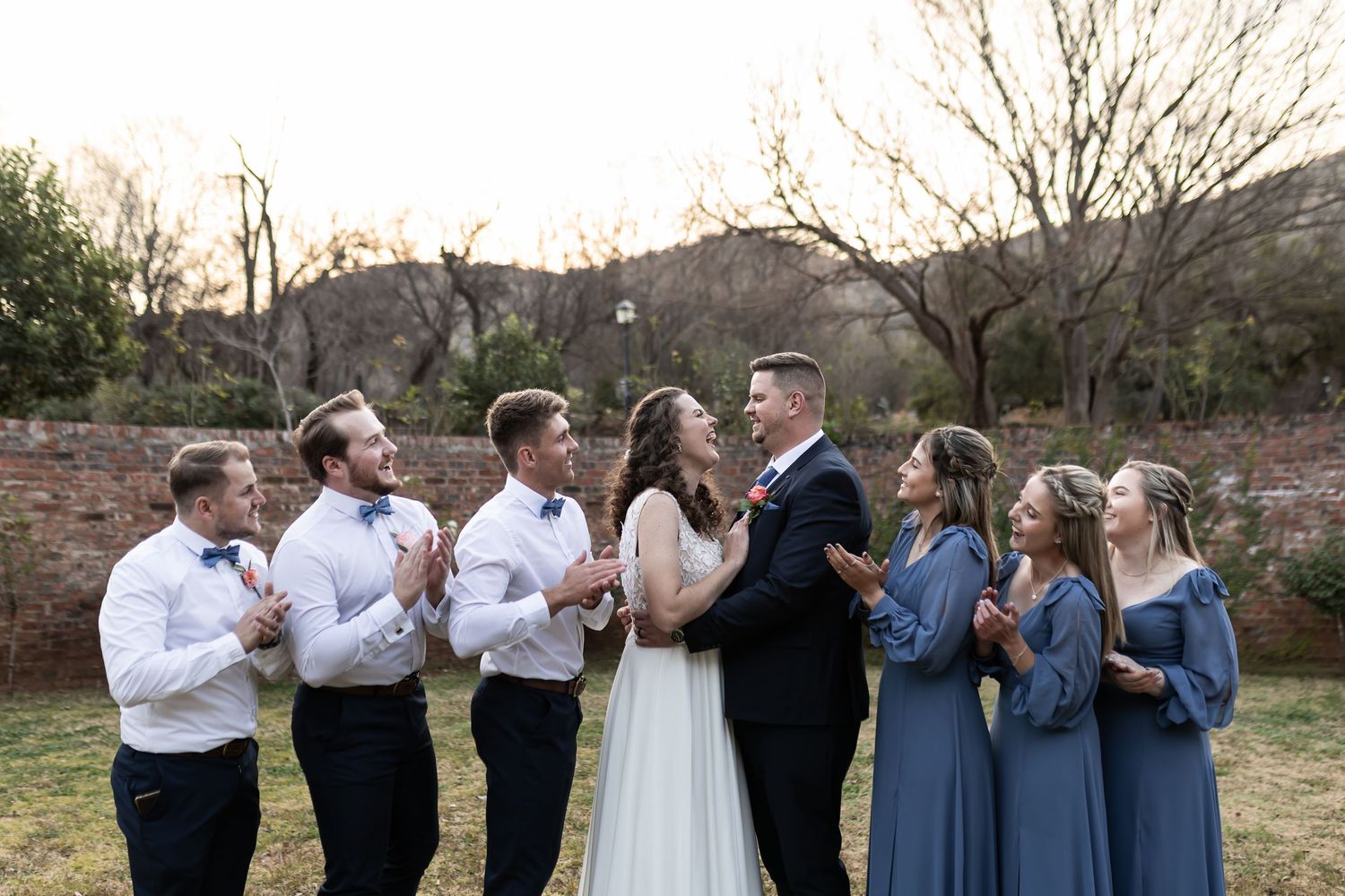 Bridal party in dusty blue dresses and white shirts posed together outdoors at sunset with desert landscape behind them.