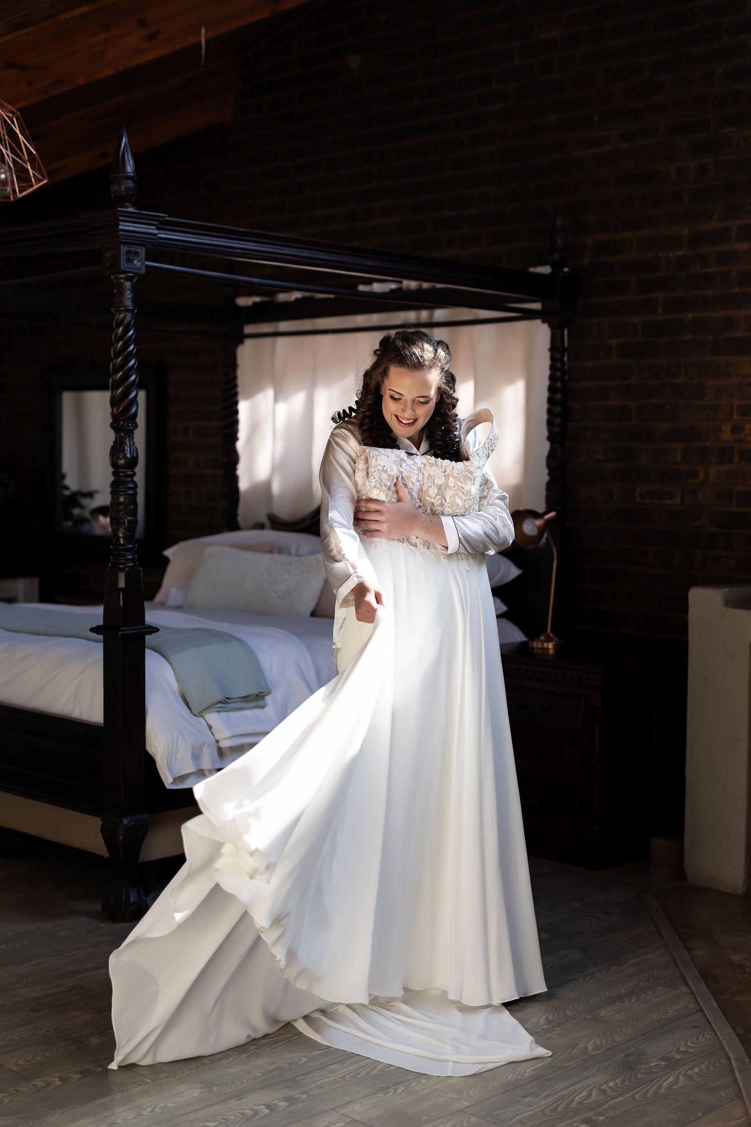 A flowing white wedding gown with long sleeves and lace details is showcased in a rustic bedroom with a canopy bed.