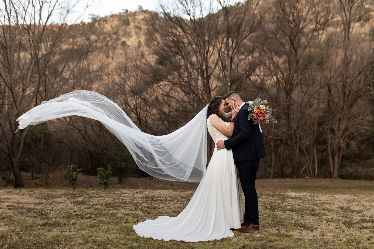 A couple poses outdoors in wedding attire with a flowing veil catching the wind against a scenic mountain backdrop.