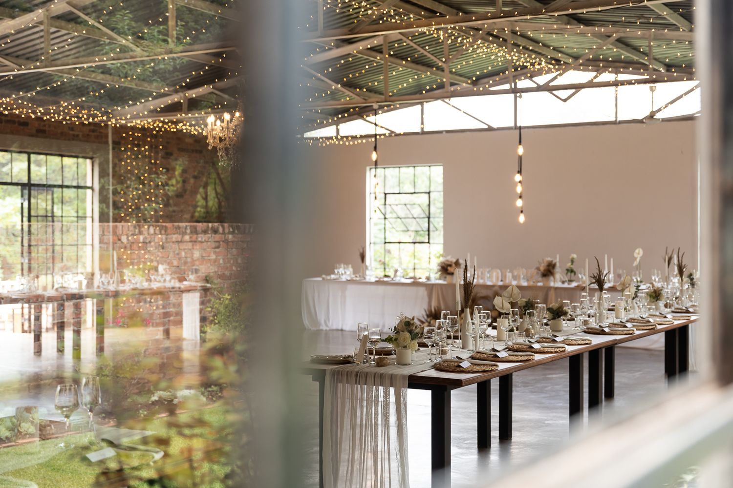 Rustic wedding reception space with industrial-style tables and hanging lights viewed through greenery.