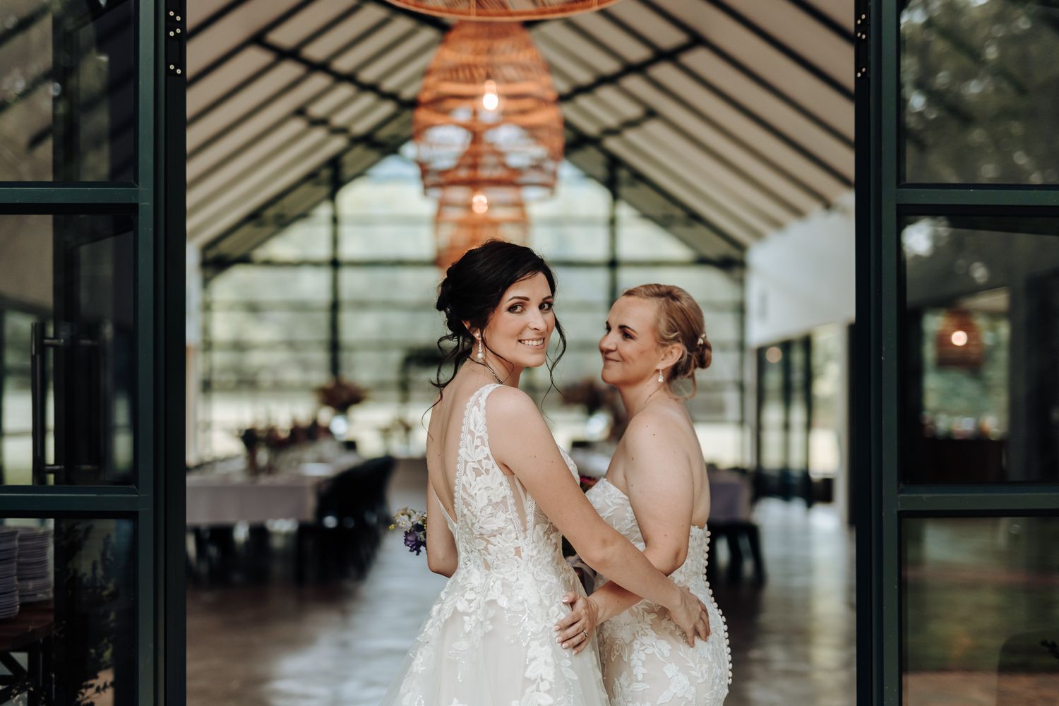 Two brides in white lace dresses pose together in front of a modern glass venue entrance.