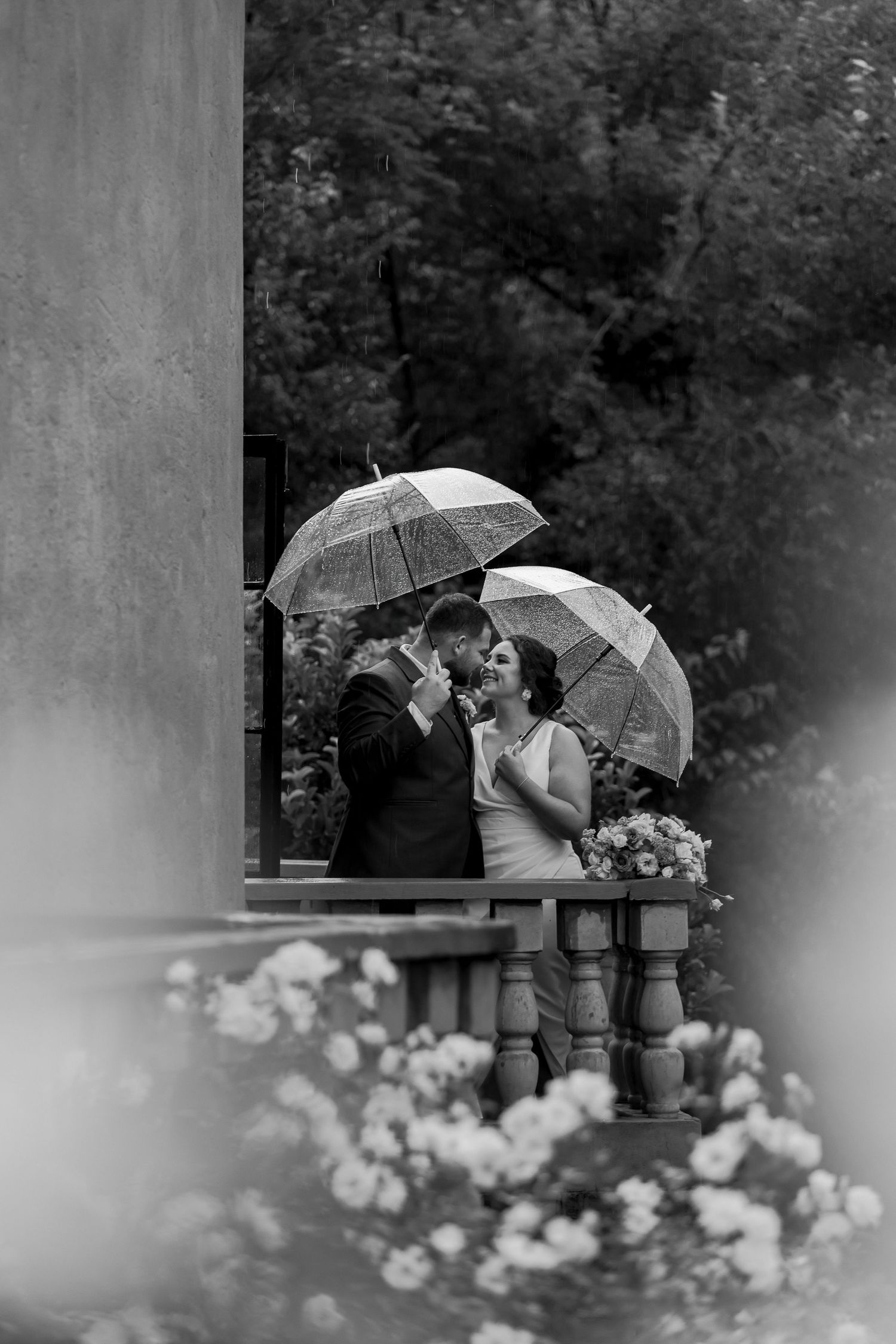 Black and white photo of newlyweds holding clear umbrellas on a balcony with white flowers below.