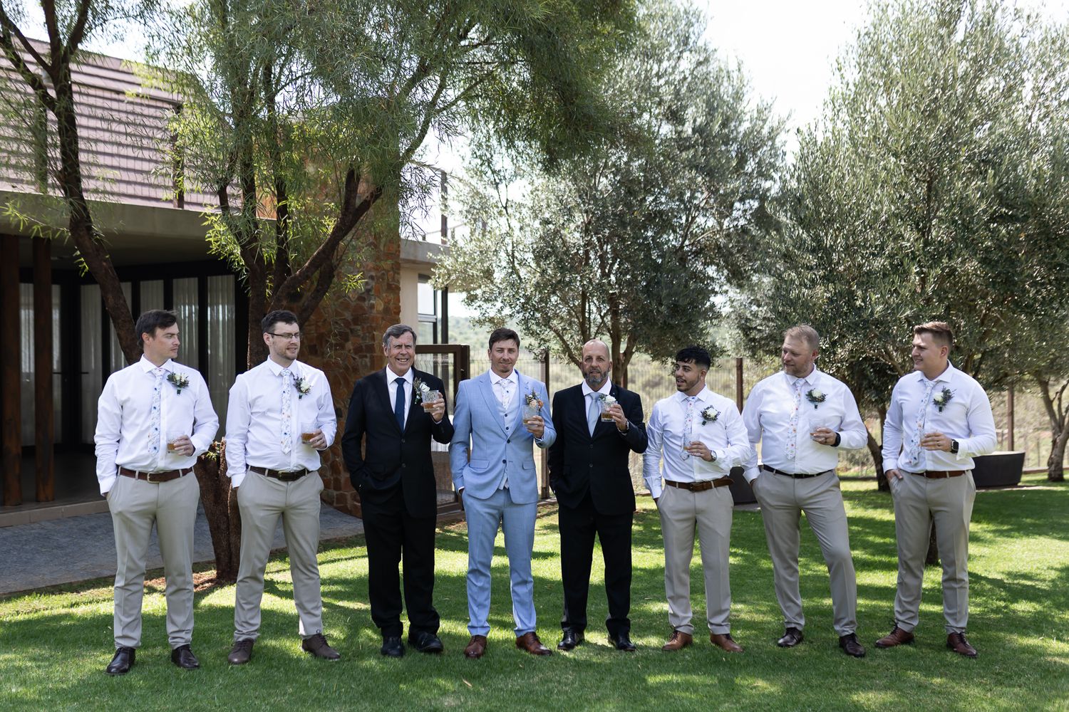 Groomsmen in light suits and one in blue stand together outdoors under olive trees.