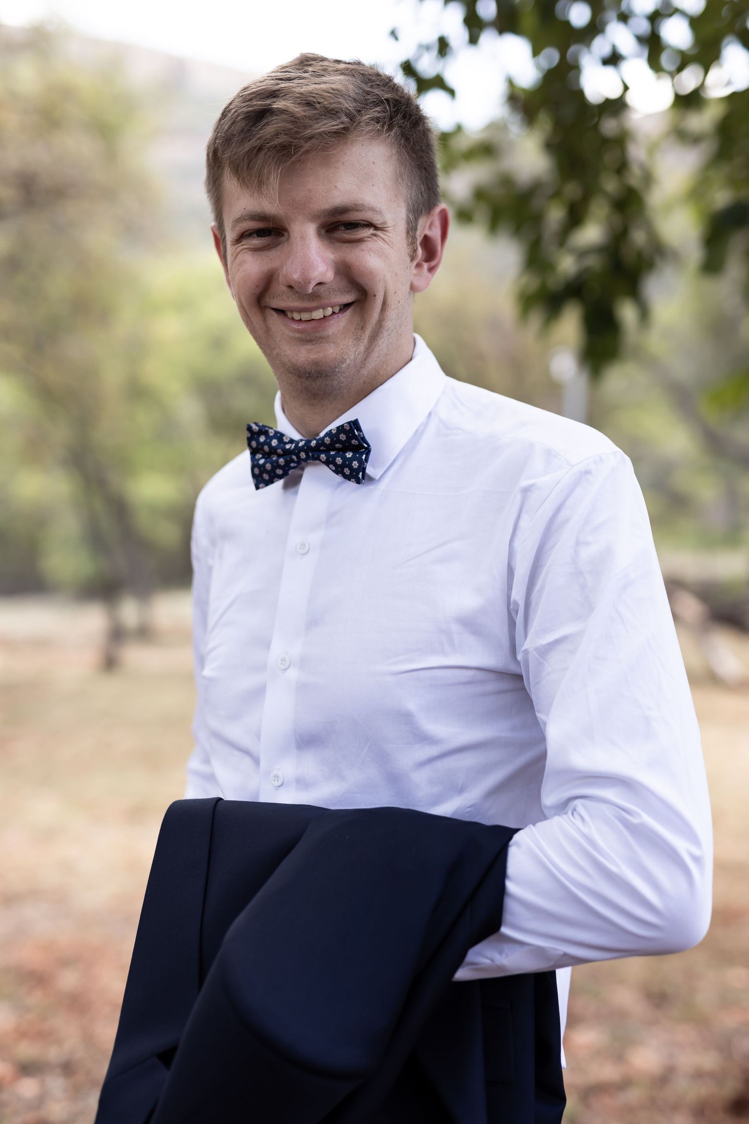 A smiling groom wearing a polka dot bow tie and white dress shirt holds his navy jacket.