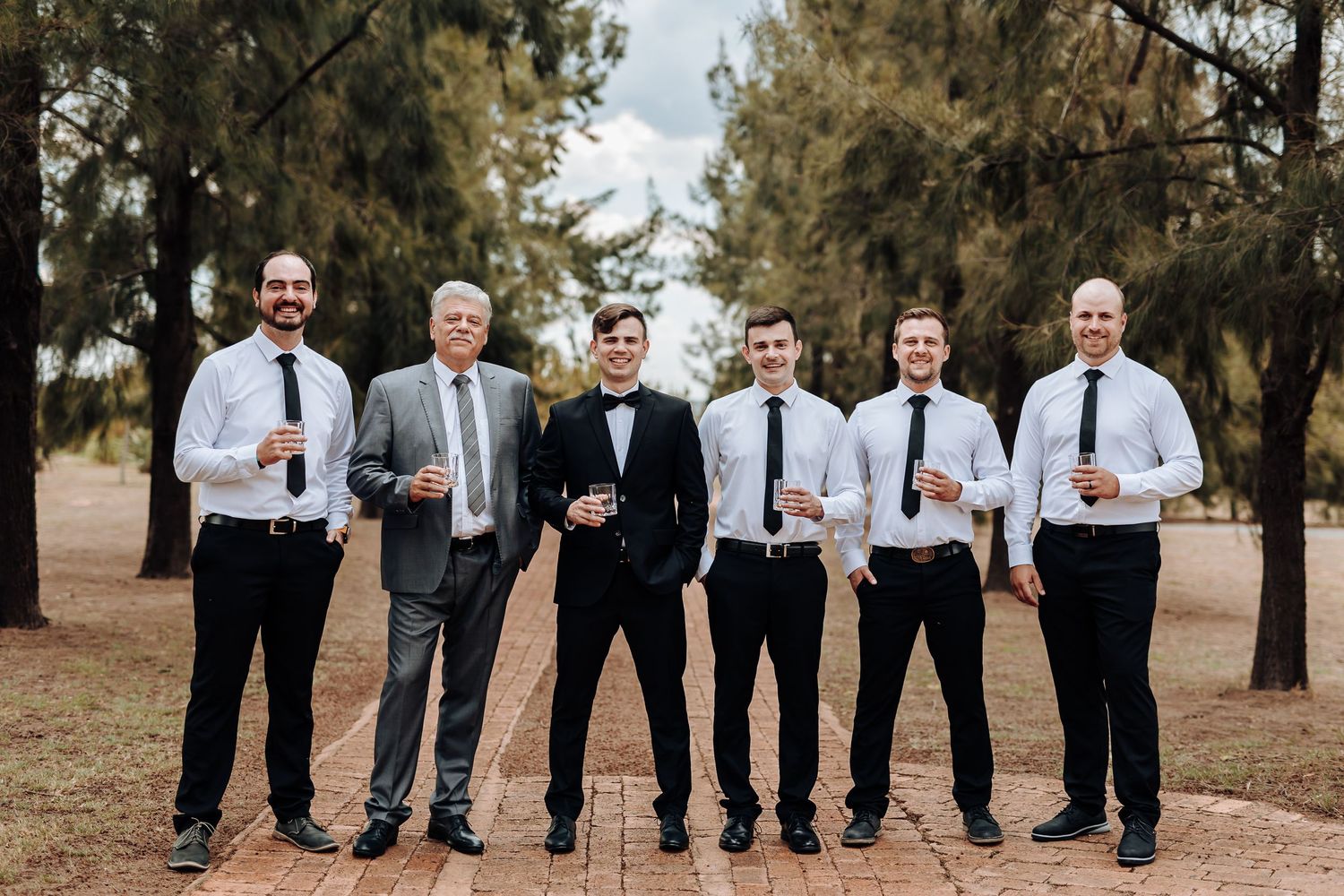 A group of groomsmen in formal attire pose together for a photo in a pine forest setting.