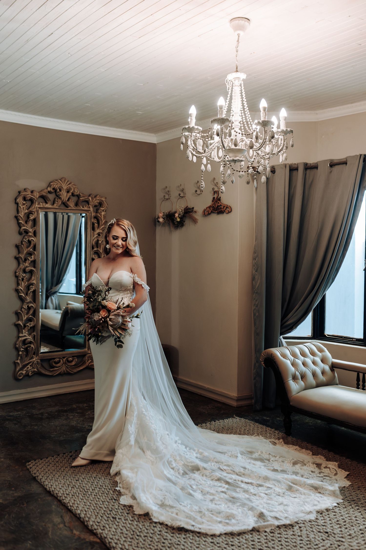 Full-length bridal portrait with flowing train under crystal chandelier in elegant room setting.