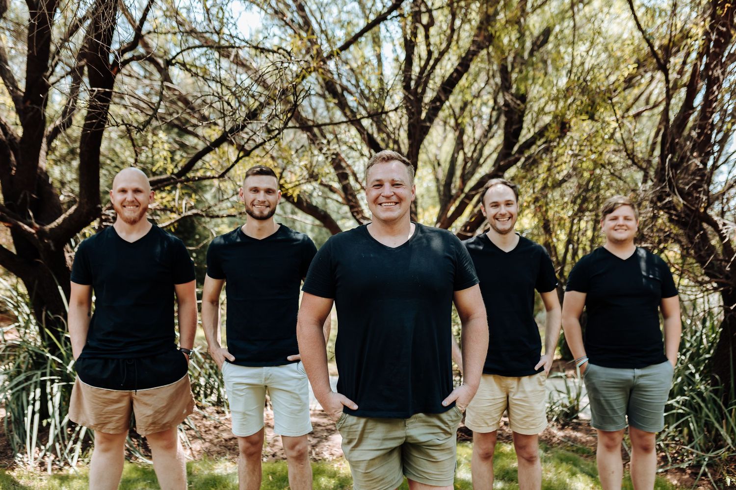 Group of men in matching black t-shirts and shorts pose for casual outdoor photo.