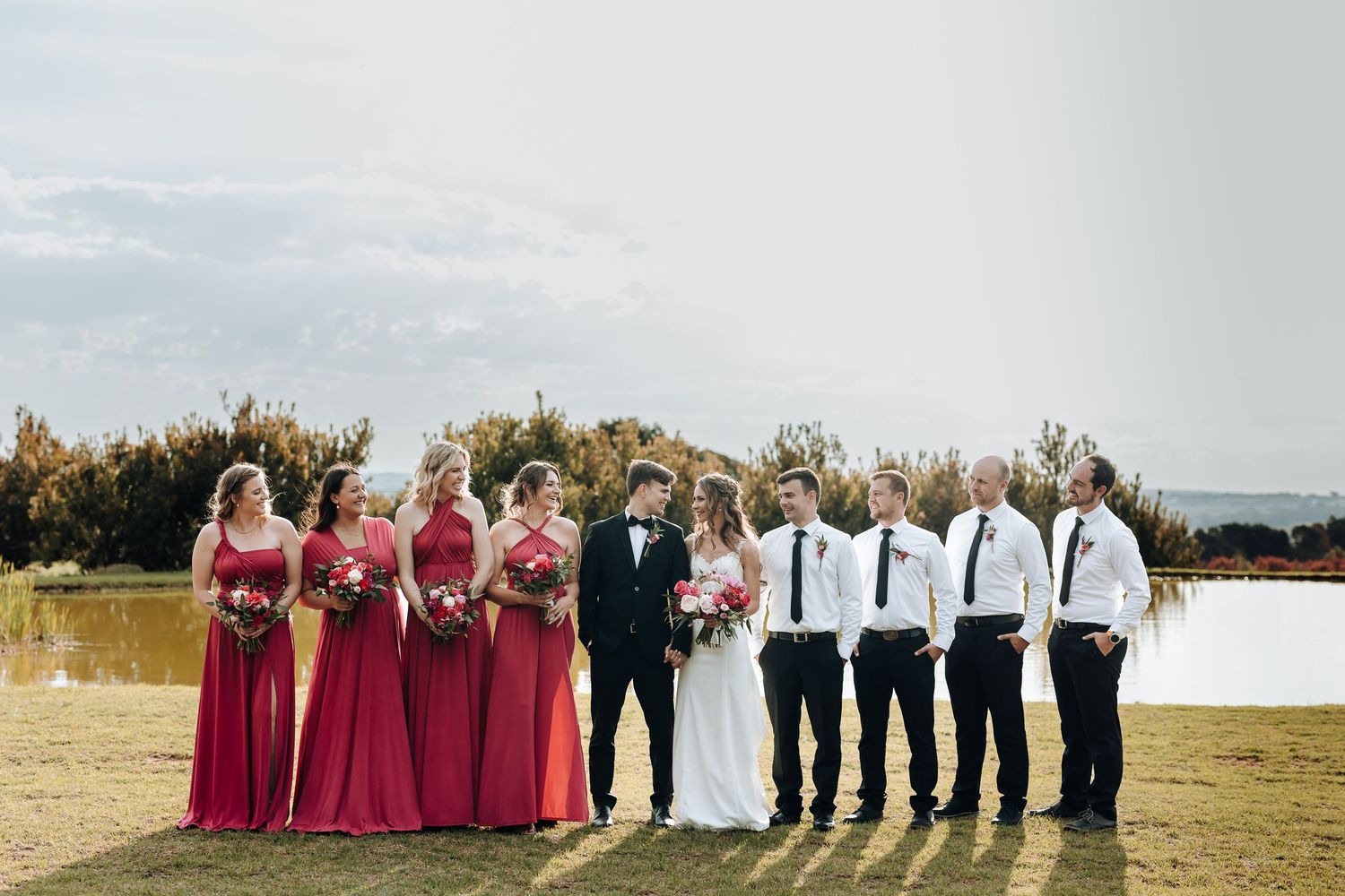 Wedding party in burgundy dresses and black tuxedo poses outdoors by a lake.
