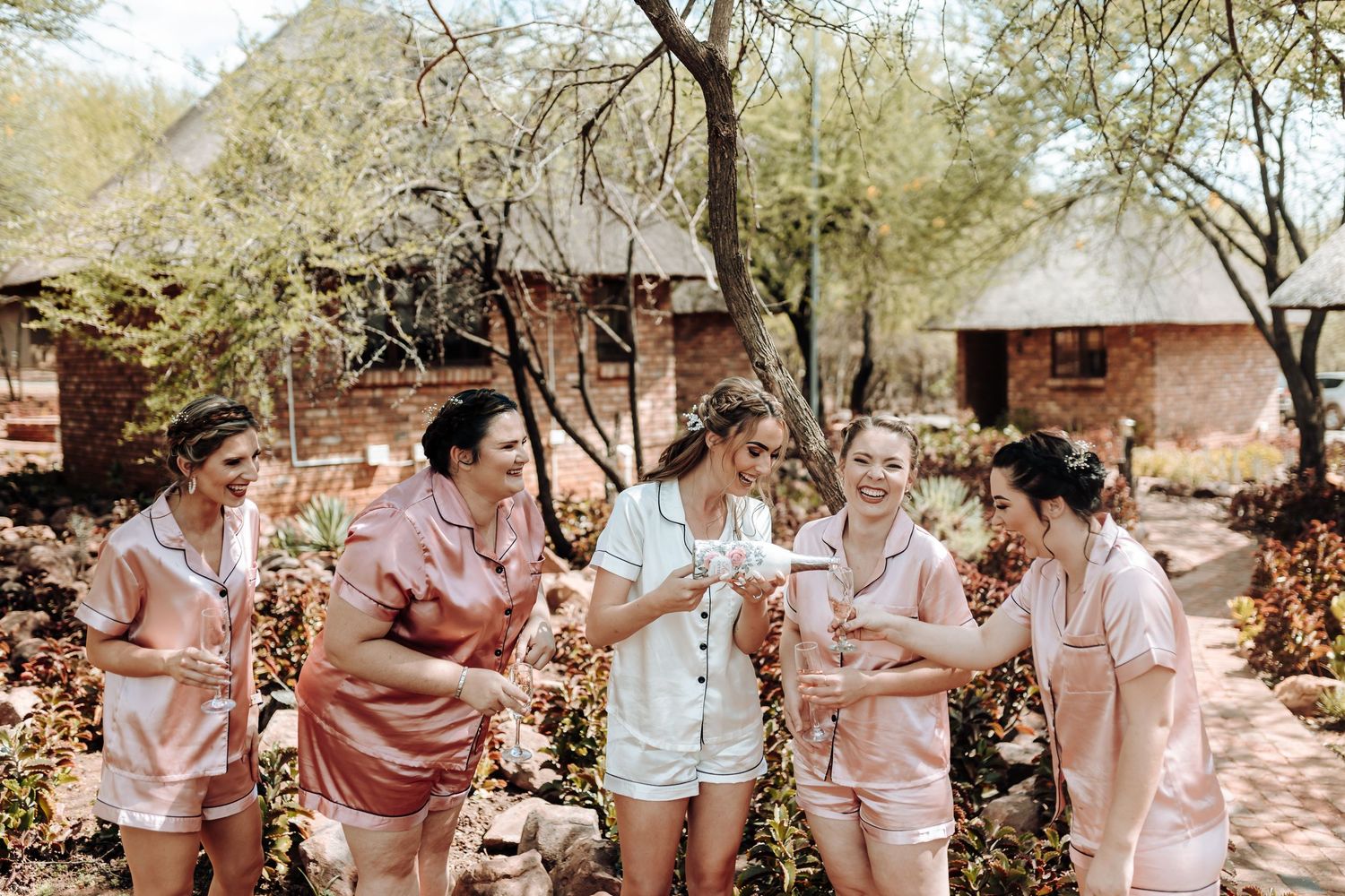Friends in pink and white satin loungewear sharing celebratory drinks in a garden.
