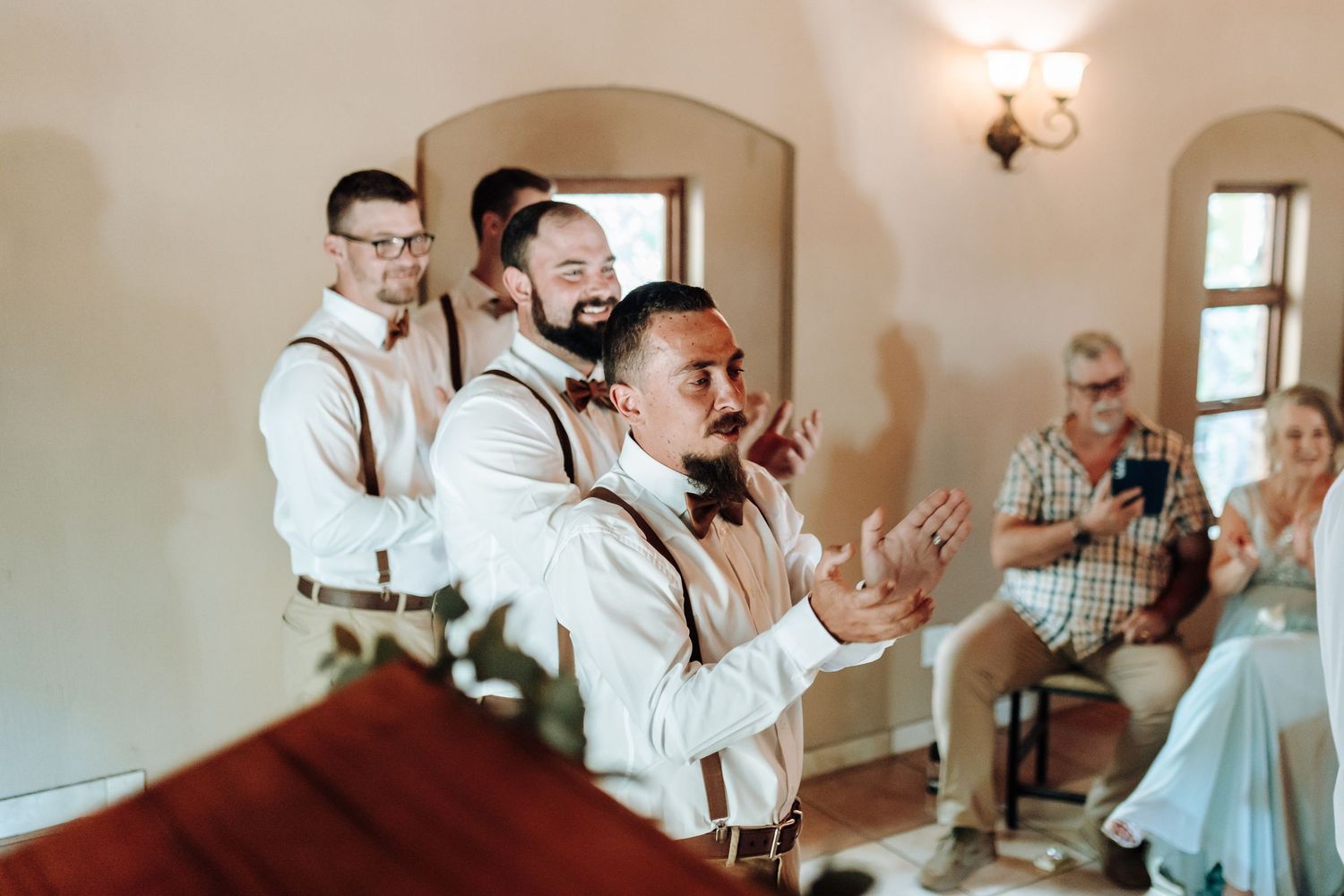 Celebration at wedding ceremony with groomsmen in white shirts and suspenders showing emotional reactions.