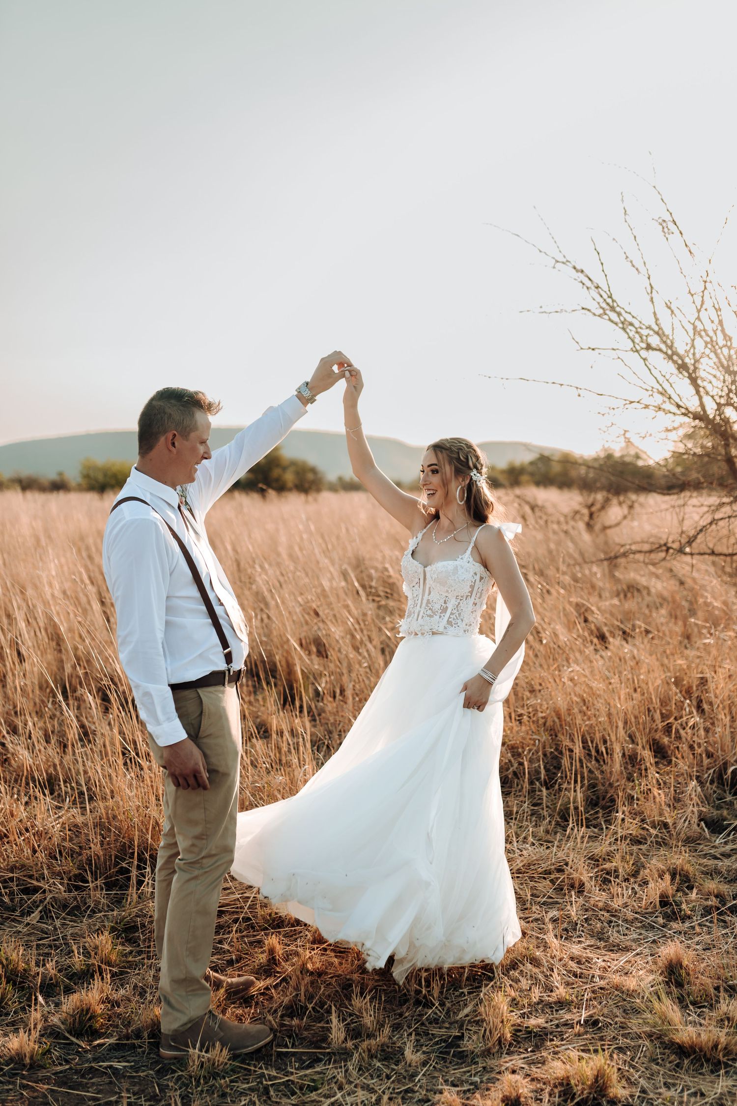 Playful wedding couple dances in rustic field with mountain backdrop at sunset.