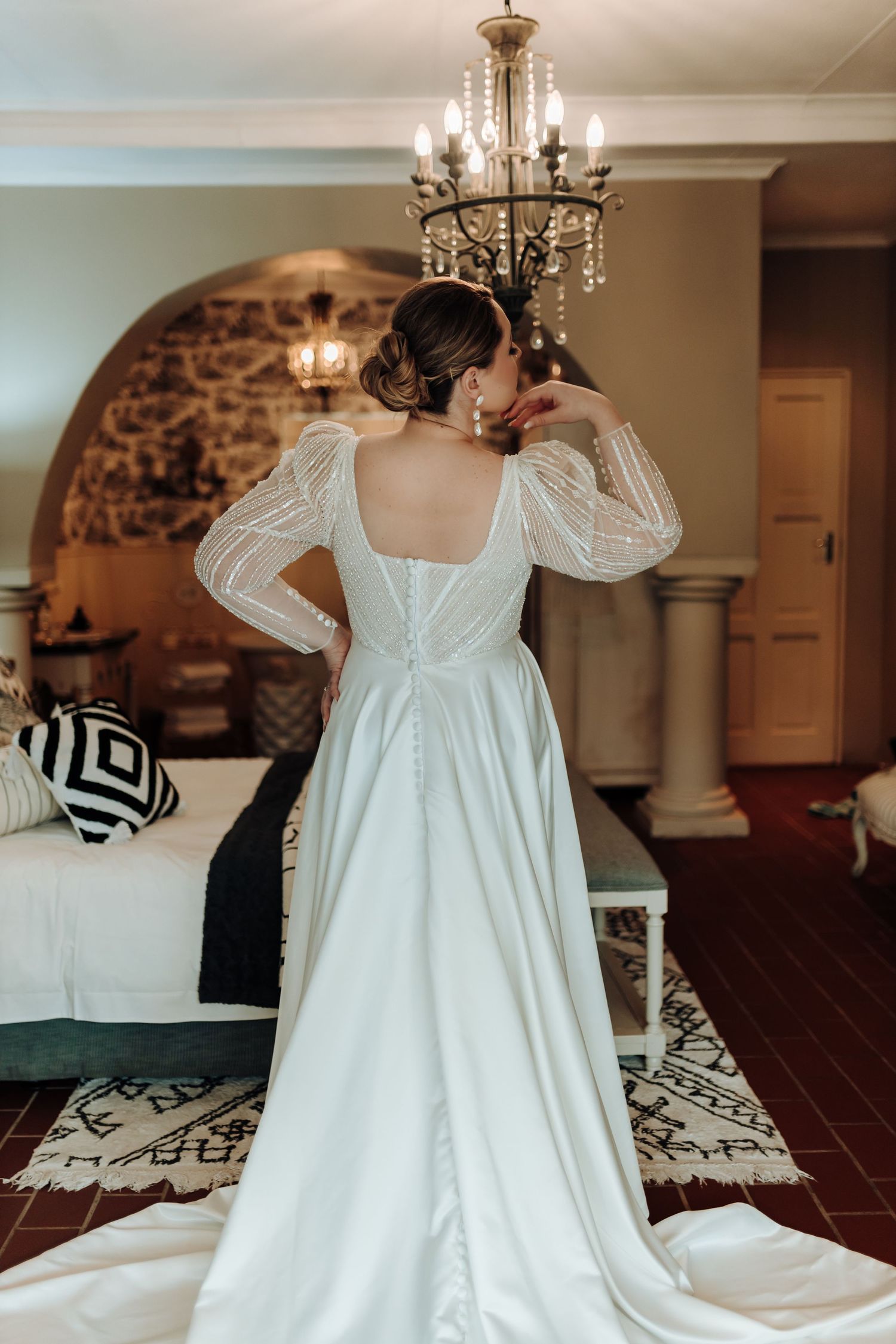 Back view of wedding dress showing intricate button details and flowing train under crystal chandelier.