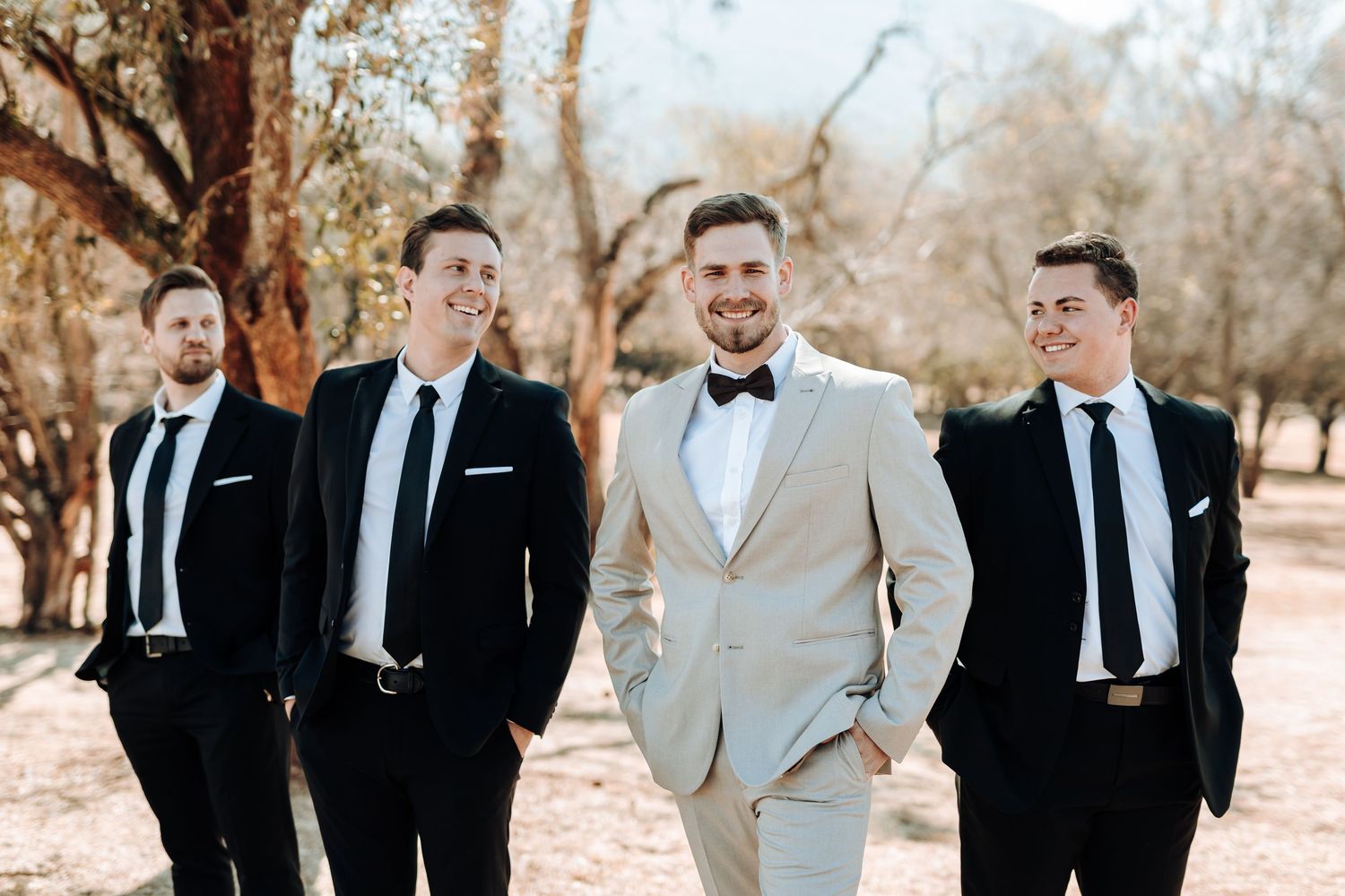 Groom in light suit poses with groomsmen in dark suits outdoors.