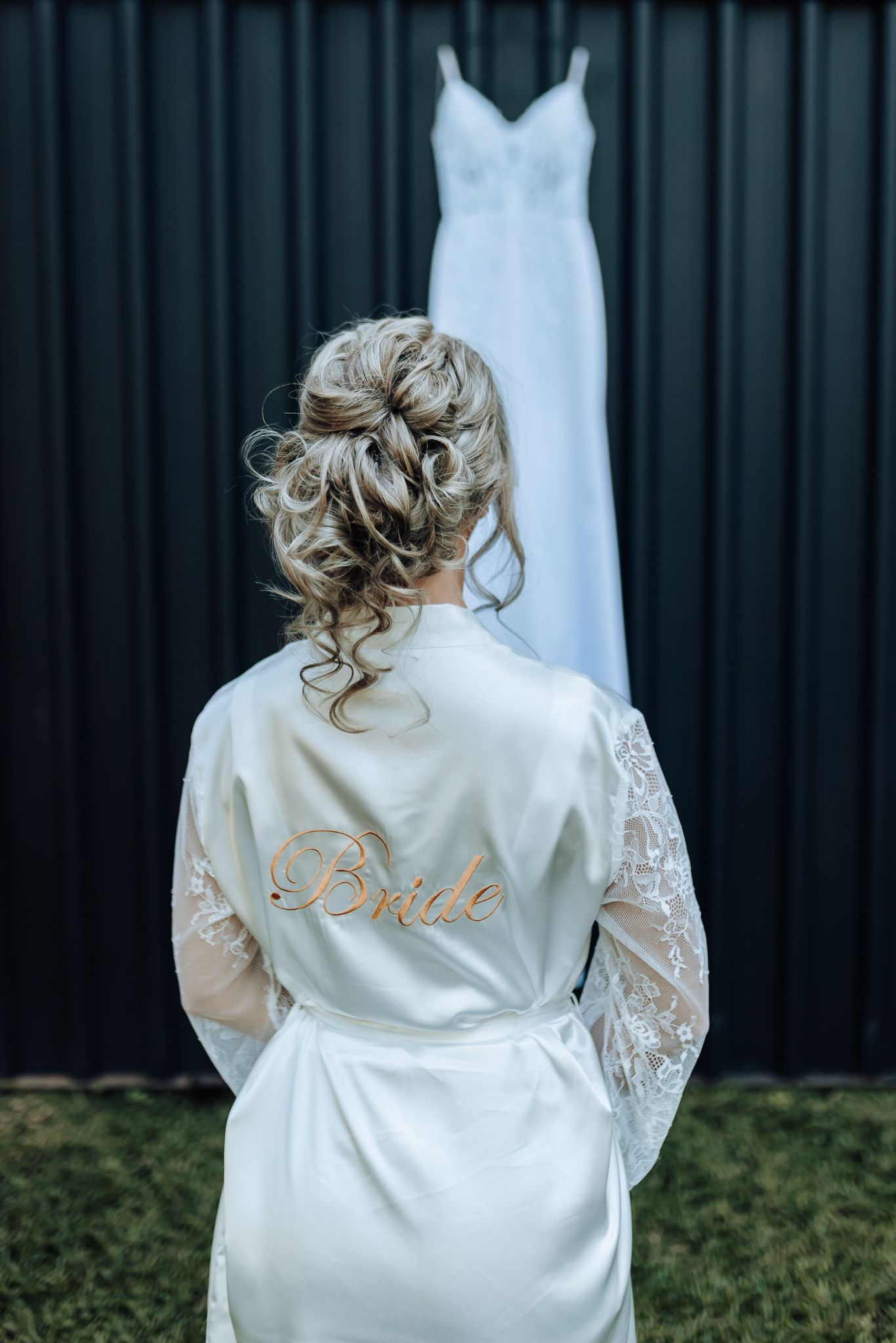 White bridal robe with lace detail and wedding dress hanging in background.