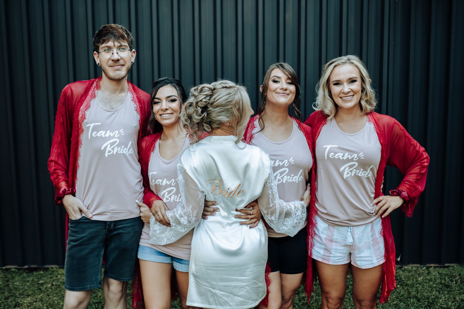 Wedding party in matching red robes and gray shirts posing against dark wall.