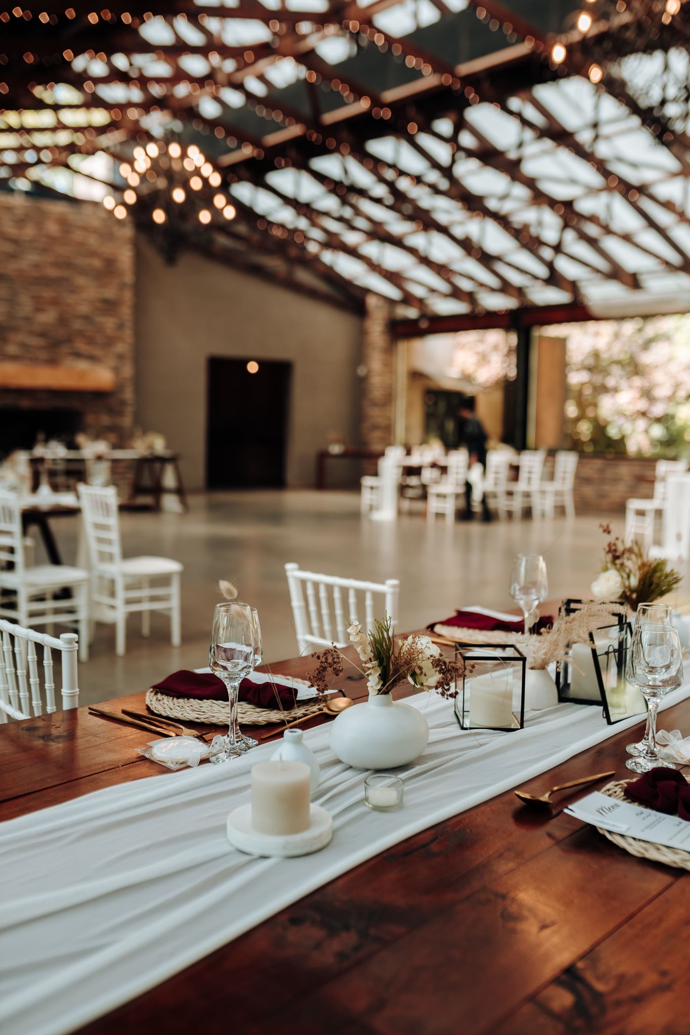 Wedding reception table decorated with white runner, burgundy accents and string lights overhead.