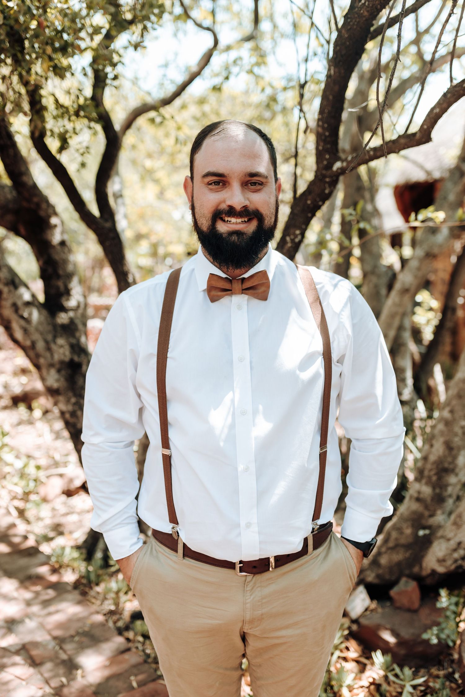 Groomsman in rustic wedding attire poses with hands in pockets among trees.