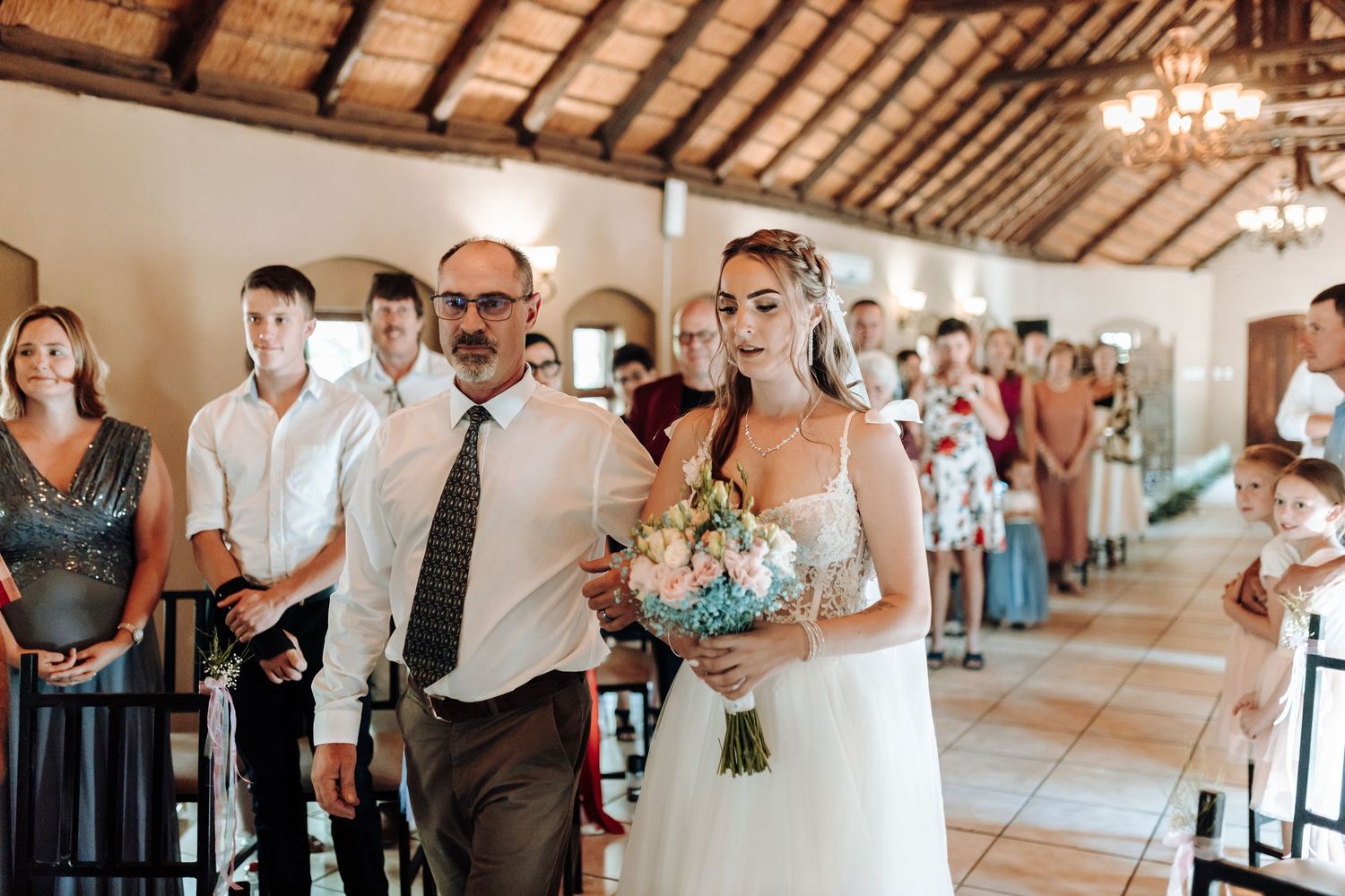 Wedding ceremony processional in a rustic venue with wooden beam ceilings and guests lining the aisle.