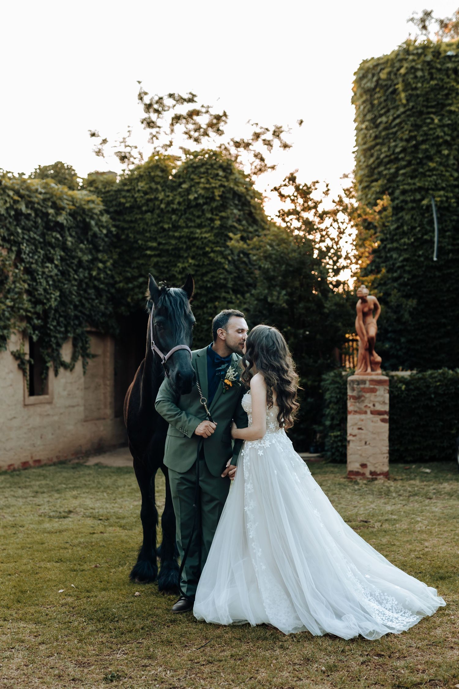 Wedding couple posing with a black horse against ivy-covered walls at sunset.
