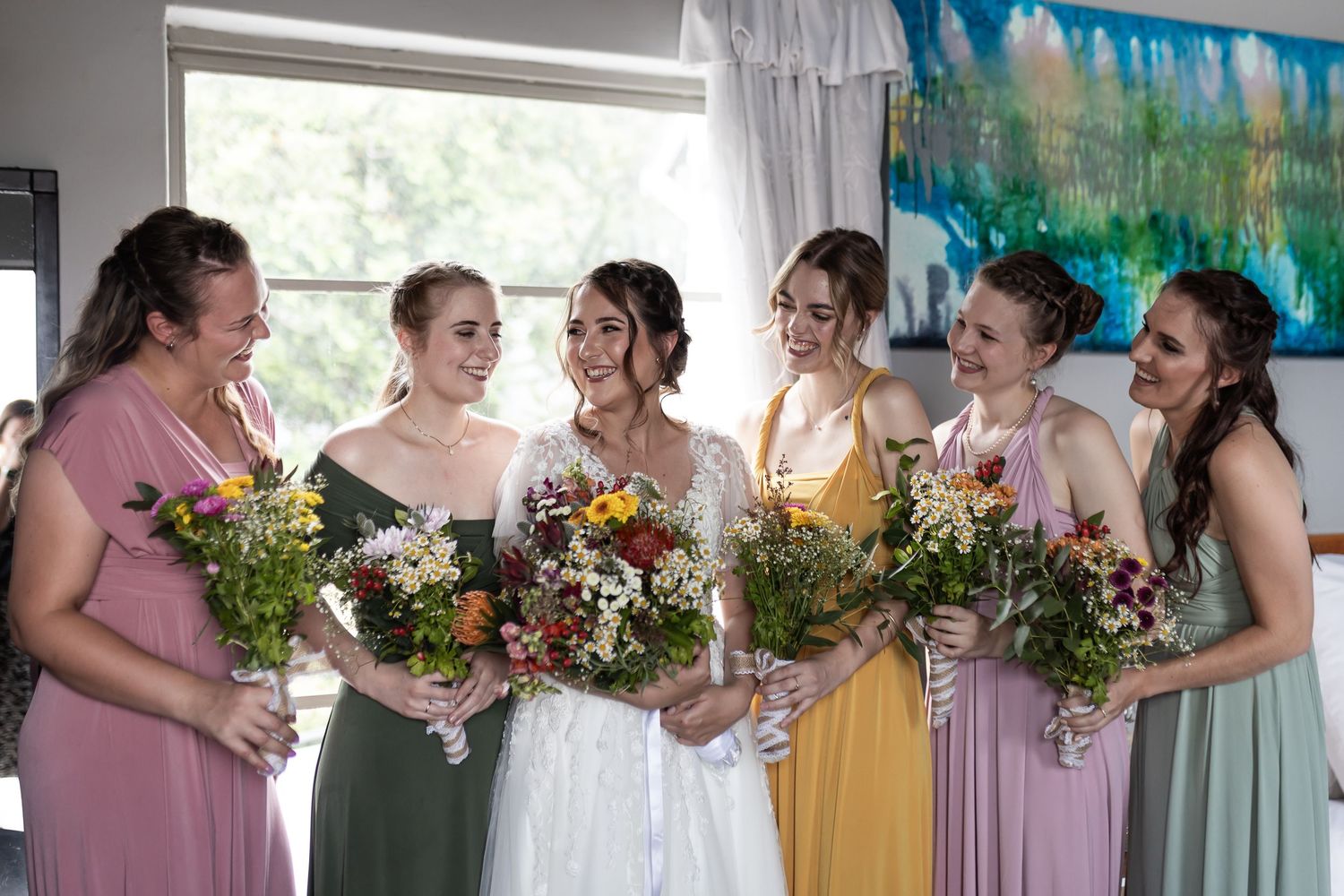 Bridesmaids holding colorful bouquets gather around the bride in a bright room.