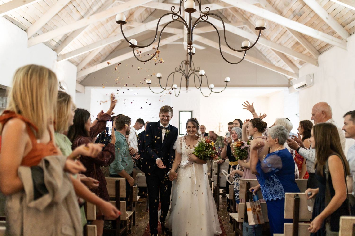 Wedding guests throw flower petals inside a bright chapel with vaulted ceilings and chandeliers.