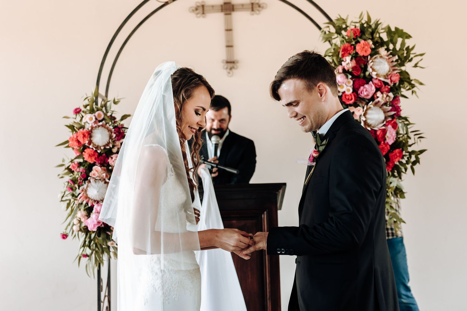 Wedding ceremony under a floral arch with pink and white roses and a cross.