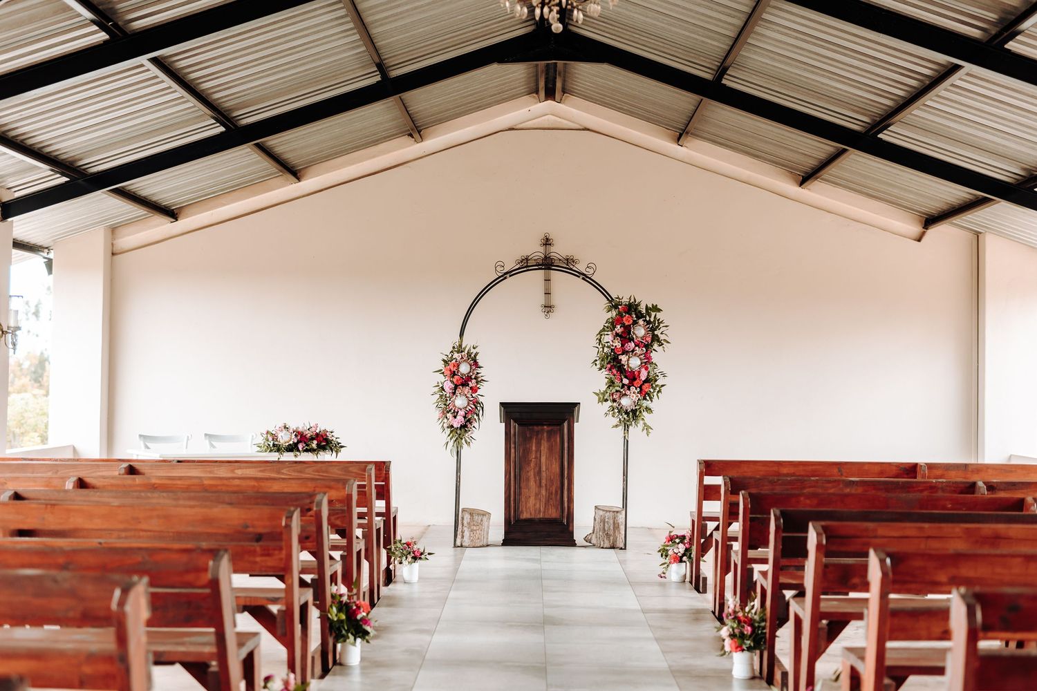 Simple white chapel interior with wooden pews and floral arch decorations at the altar.