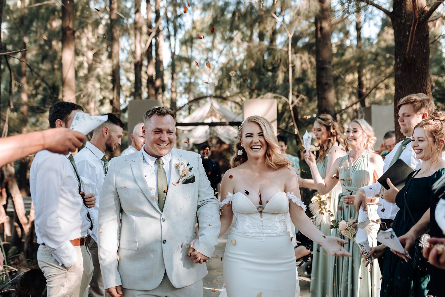 Wedding guests celebrate and throw confetti while the newlyweds walk down the aisle in an outdoor forest setting.