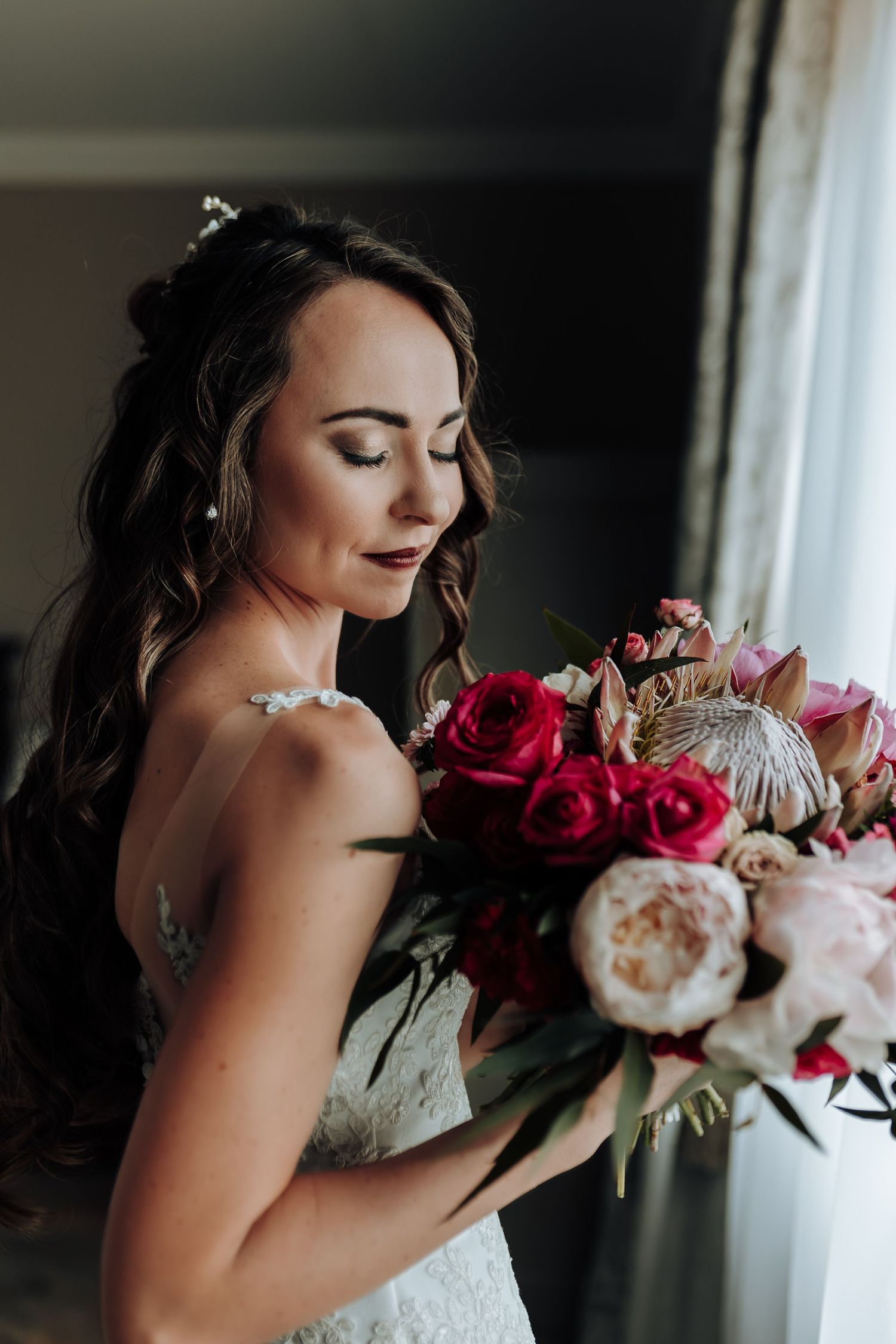 Bride in lace dress holding romantic bouquet with pink and white flowers near window.