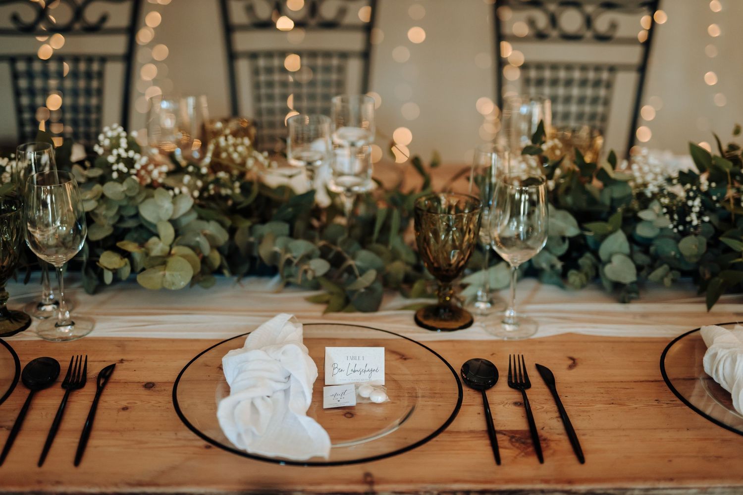 Elegant wedding table setting with black plates, green eucalyptus garland, and glowing string lights in the background.