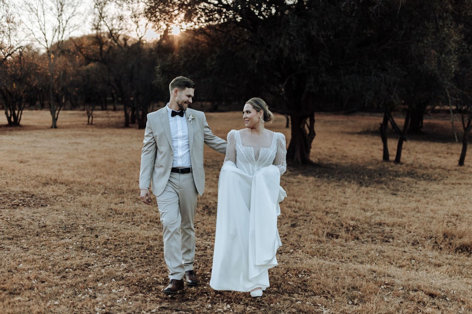 Casual wedding couple walking together through rustic field at dusk.