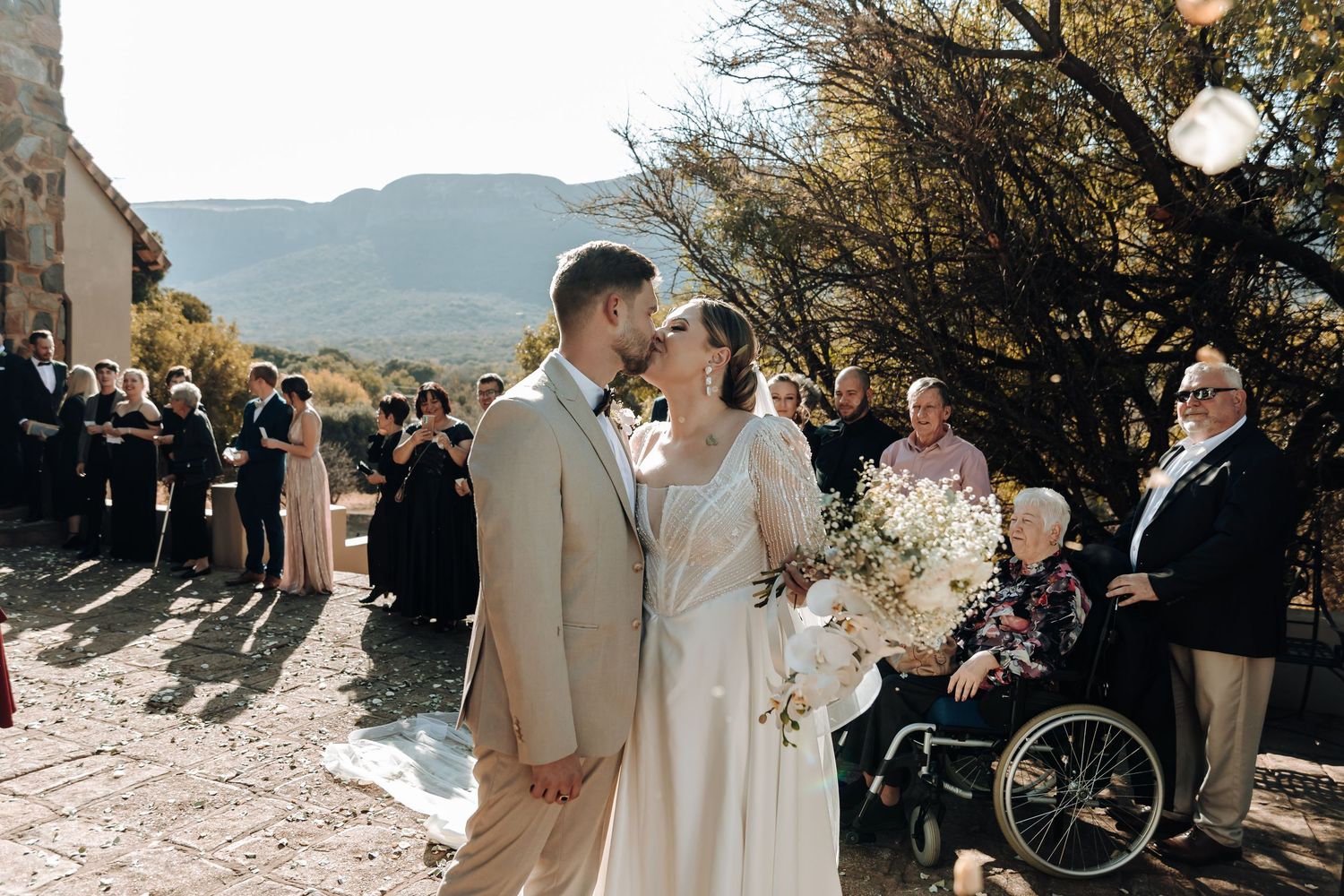 Wedding couple poses for photos outside rustic stone chapel with mountains in background.
