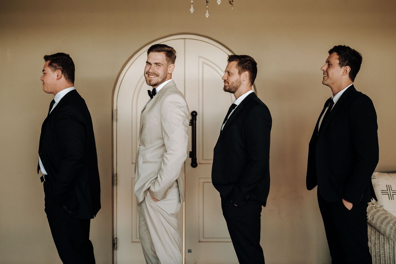 Wedding party poses in profile against white door with groom in light suit standing out from groomsmen.