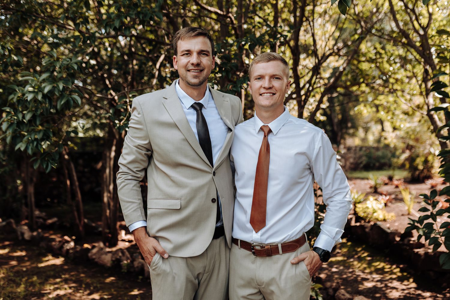 Two men in coordinating business attire pose together in outdoor woodland setting.