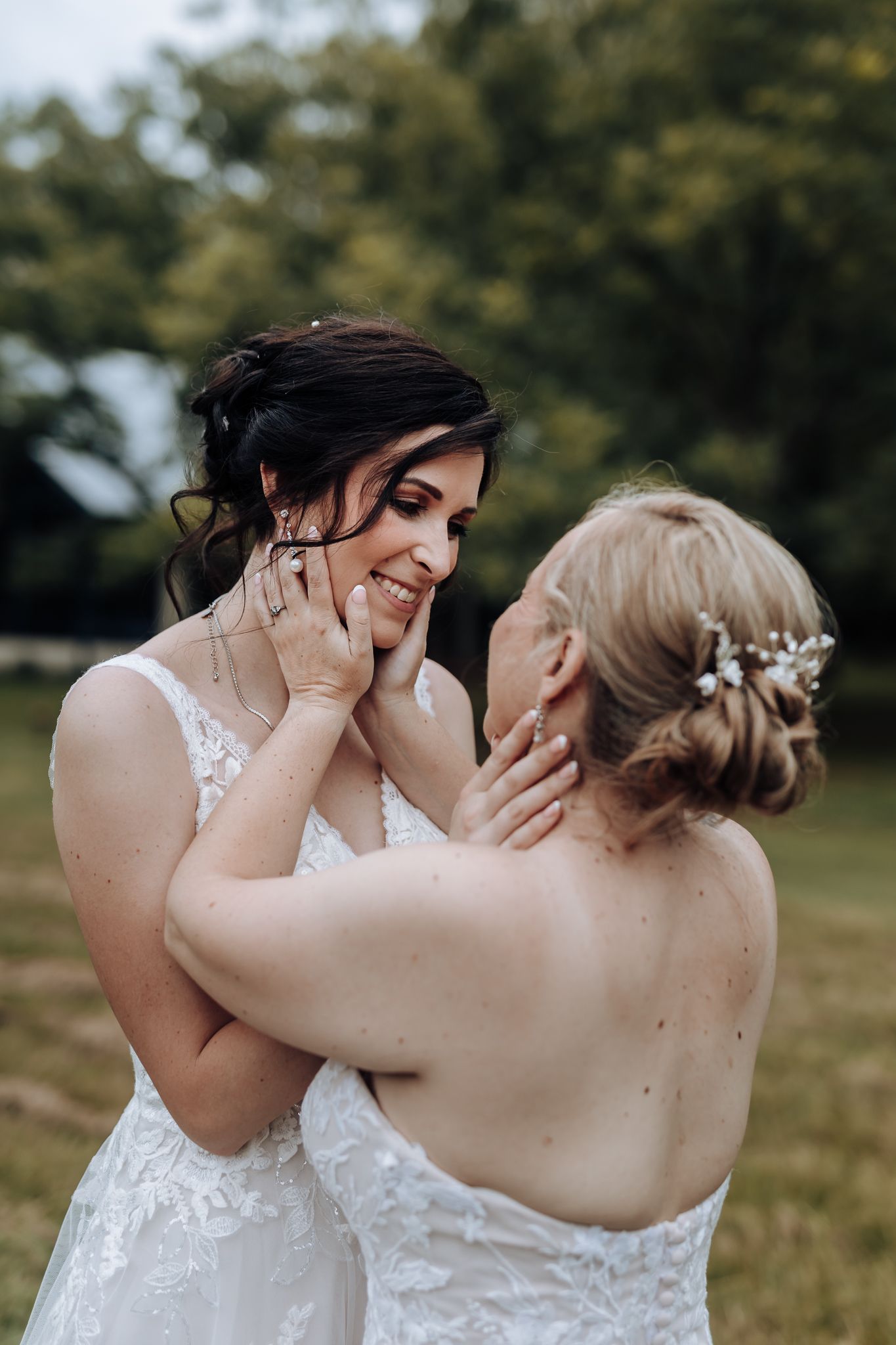 Two brides in white lace gowns tenderly hold each other's faces in an outdoor setting.