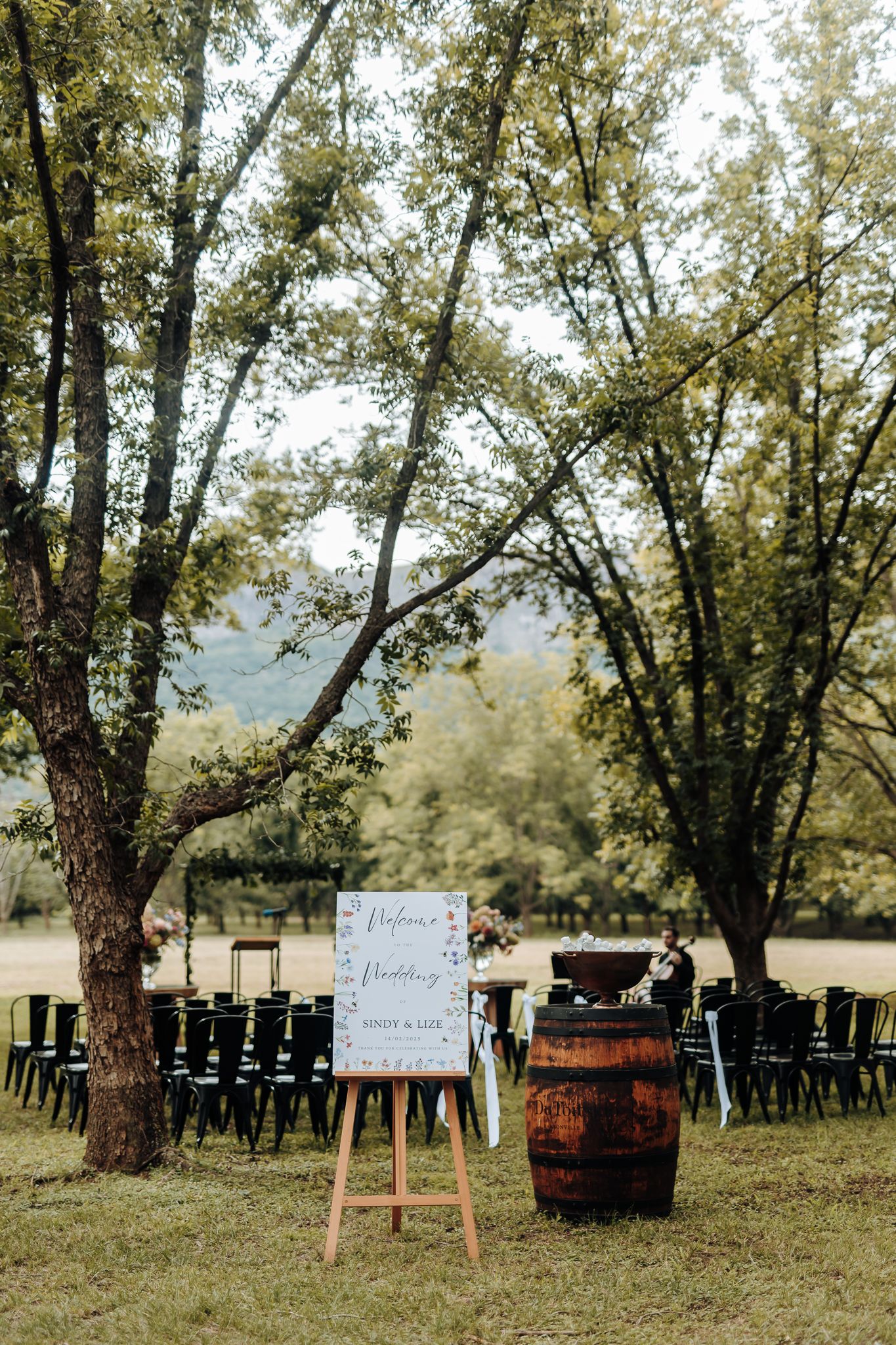 Outdoor wedding ceremony setup with rows of black chairs, a wine barrel, and a welcome sign under tall trees.