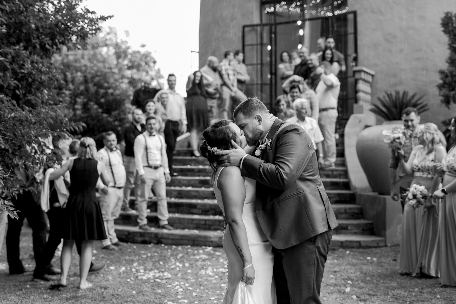 Black and white photo of wedding couple kissing outdoors surrounded by guests on stone steps.