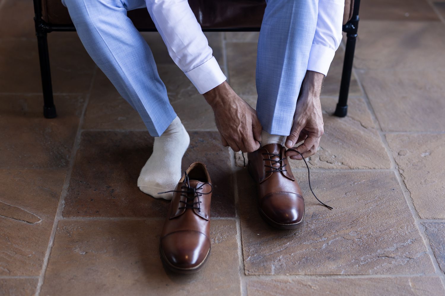 Person tying brown leather dress shoes while wearing light blue pants.