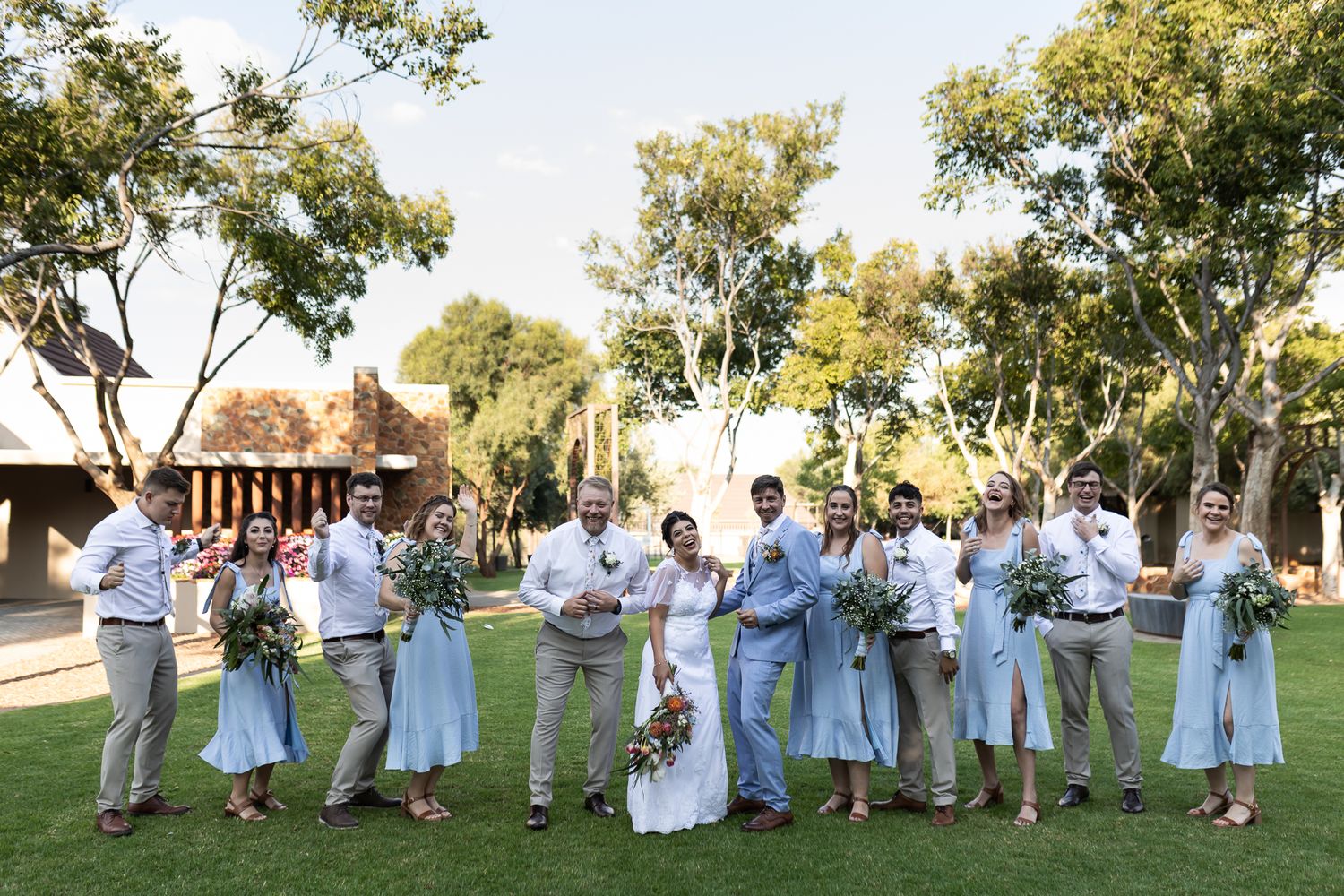 Bridal party in pastel blue dresses and khaki suits posed outdoors with bouquets.