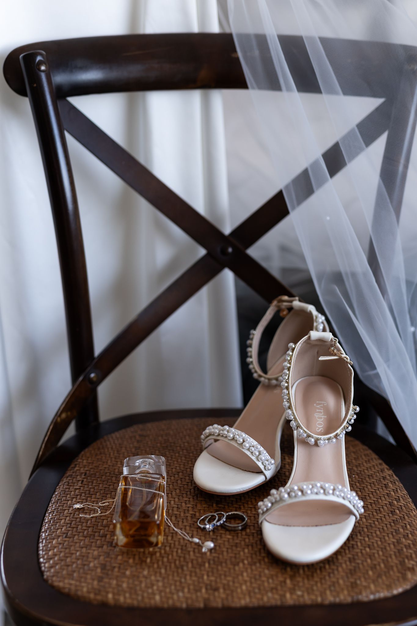 Wedding accessories including jeweled sandals and rings arranged on wooden cross-back chair.