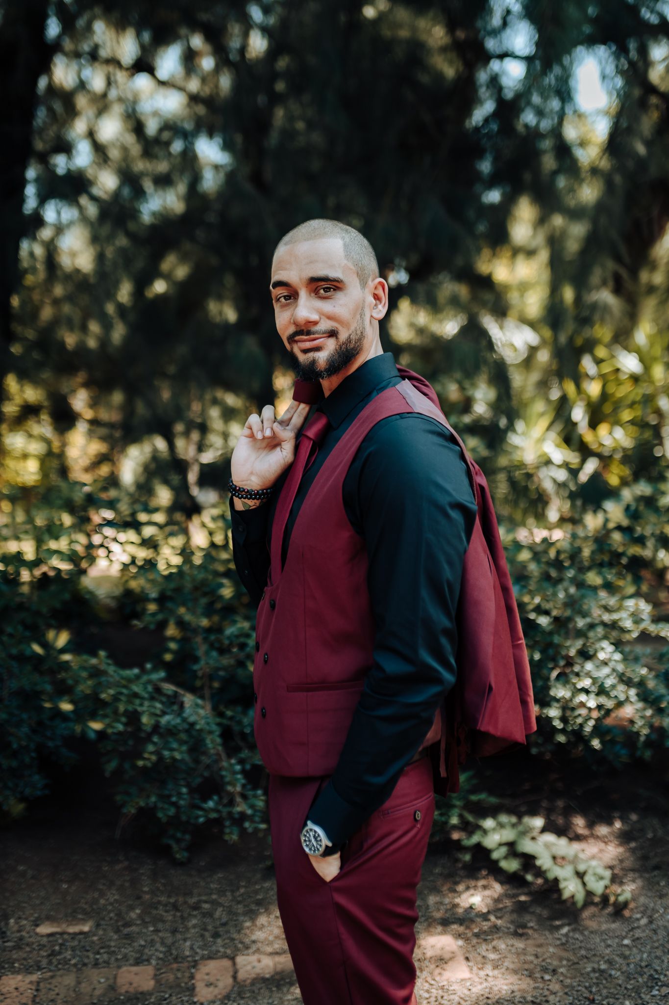 Groom in burgundy vest and black shirt poses casually in garden setting.